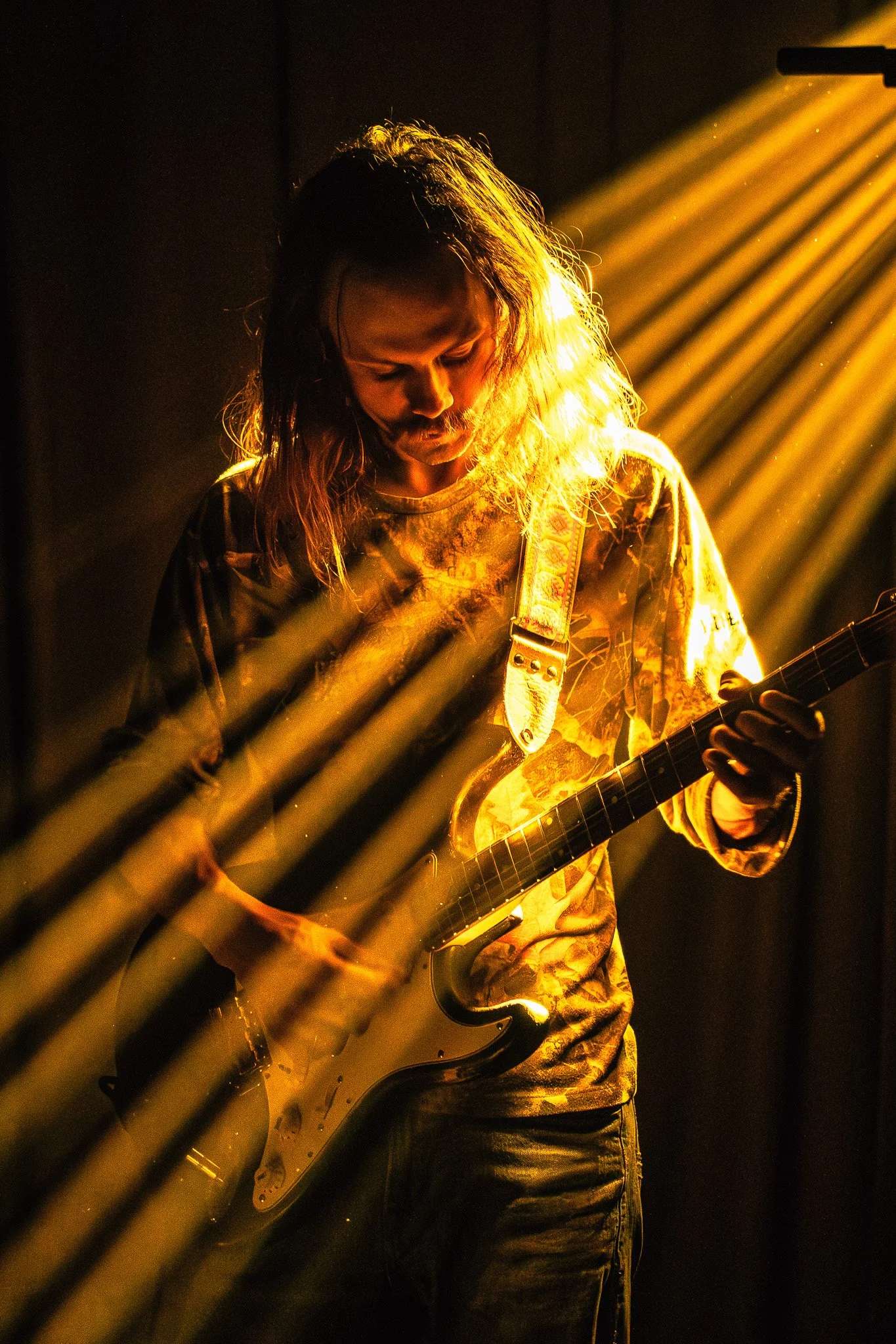 A male musician with long hair playing an electric guitar under warm, dramatic stage lighting, seen through a metal railing in the foreground.