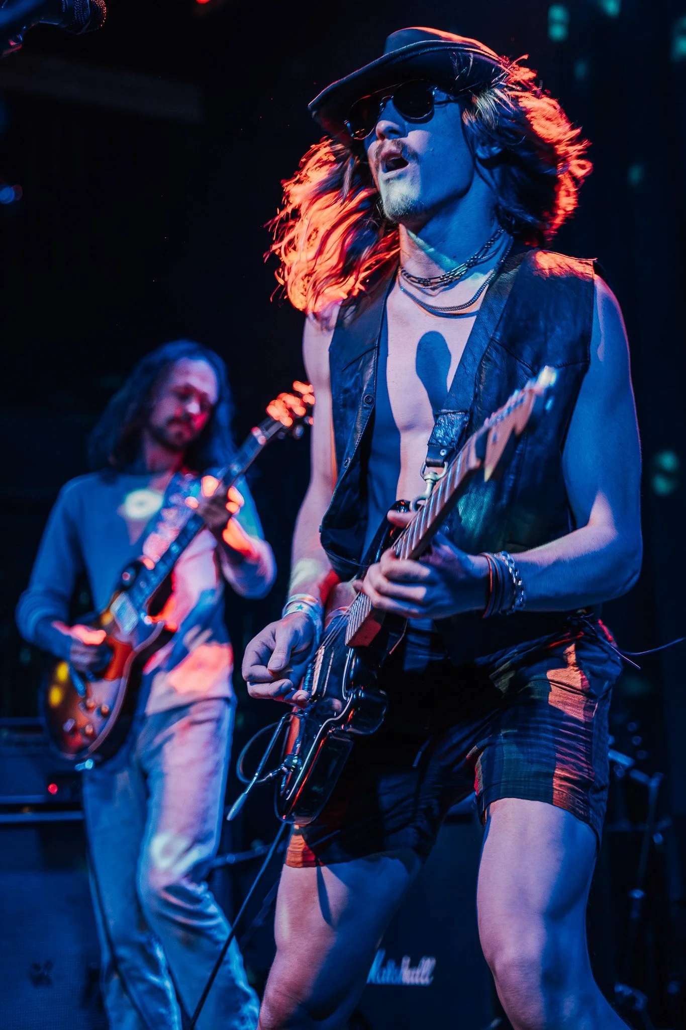 Two male musicians performing on stage with electric guitars under colorful stage lights, one with long hair, sunglasses, and a cowboy hat, the other with long hair and a beard.