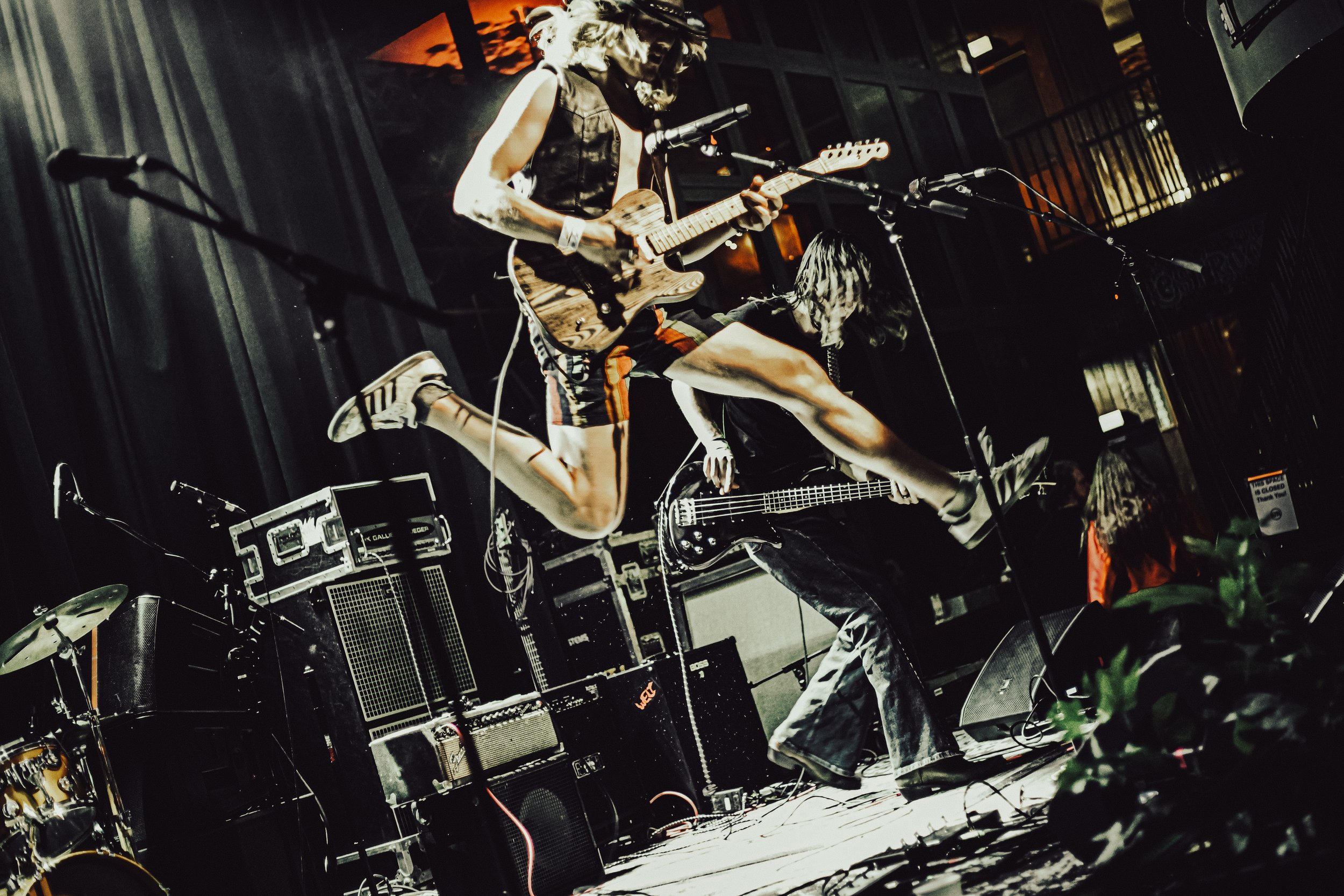 A person performing on stage at a live music event, playing an electric guitar and wearing casual clothes in a dimly lit venue with musical equipment around.