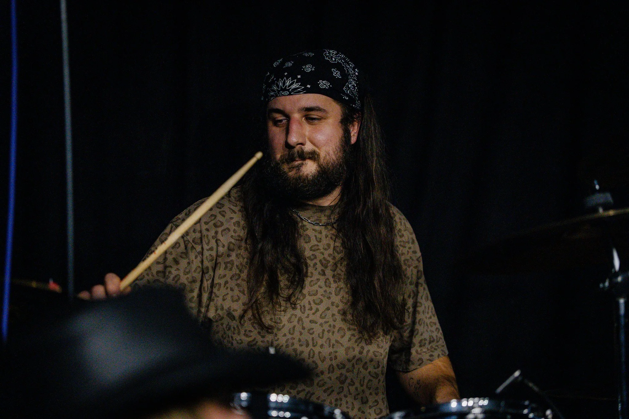 A man with long dark hair and a beard, wearing a black bandana with white patterns, playing drums in a dark room.