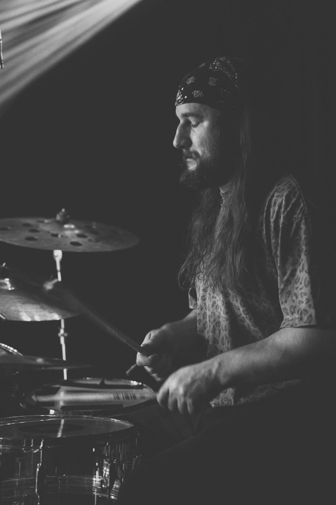 A black and white photo of a man with long hair and a bandana on his head, playing drums in a dark room.