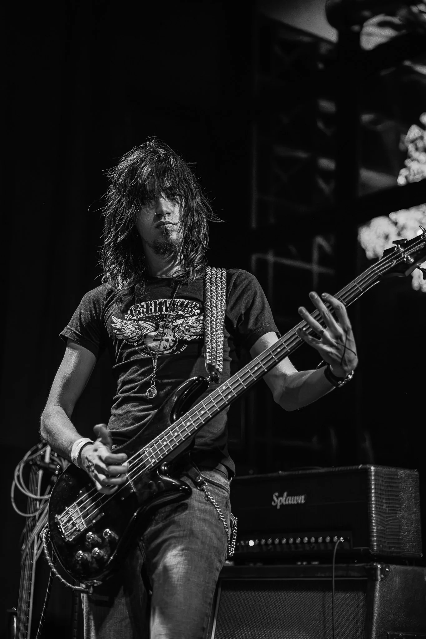 A musician with long hair and a beard playing a bass guitar on stage, with a speaker and amplifier in the background, in a black and white photograph.
