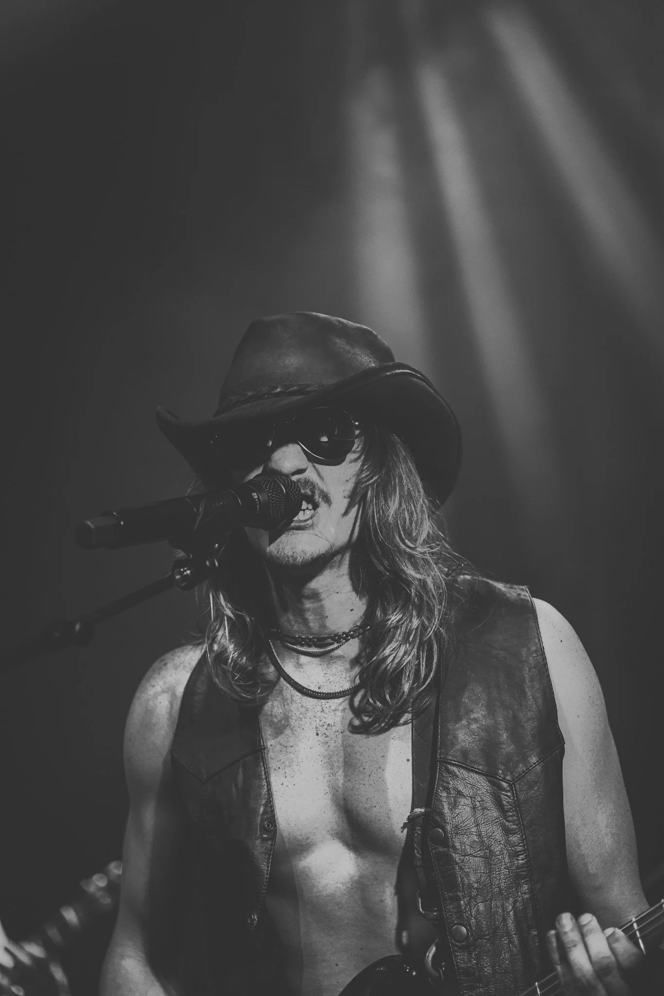 A person singing into a microphone, wearing a cowboy hat, sunglasses, a leather vest, and multiple necklaces, with long hair, in a black and white photo likely at a concert or performance.