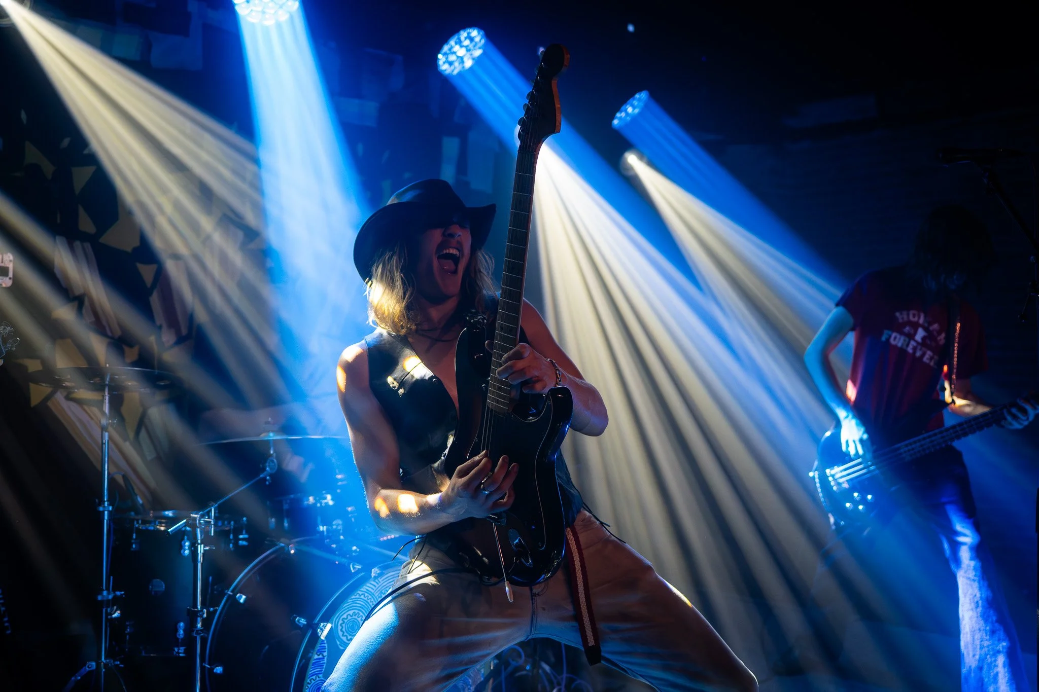 A woman wearing a cowboy hat and sunglasses playing an electric guitar on stage with blue and white stage lights shining behind her, a drummer and another guitarist are visible in the background.