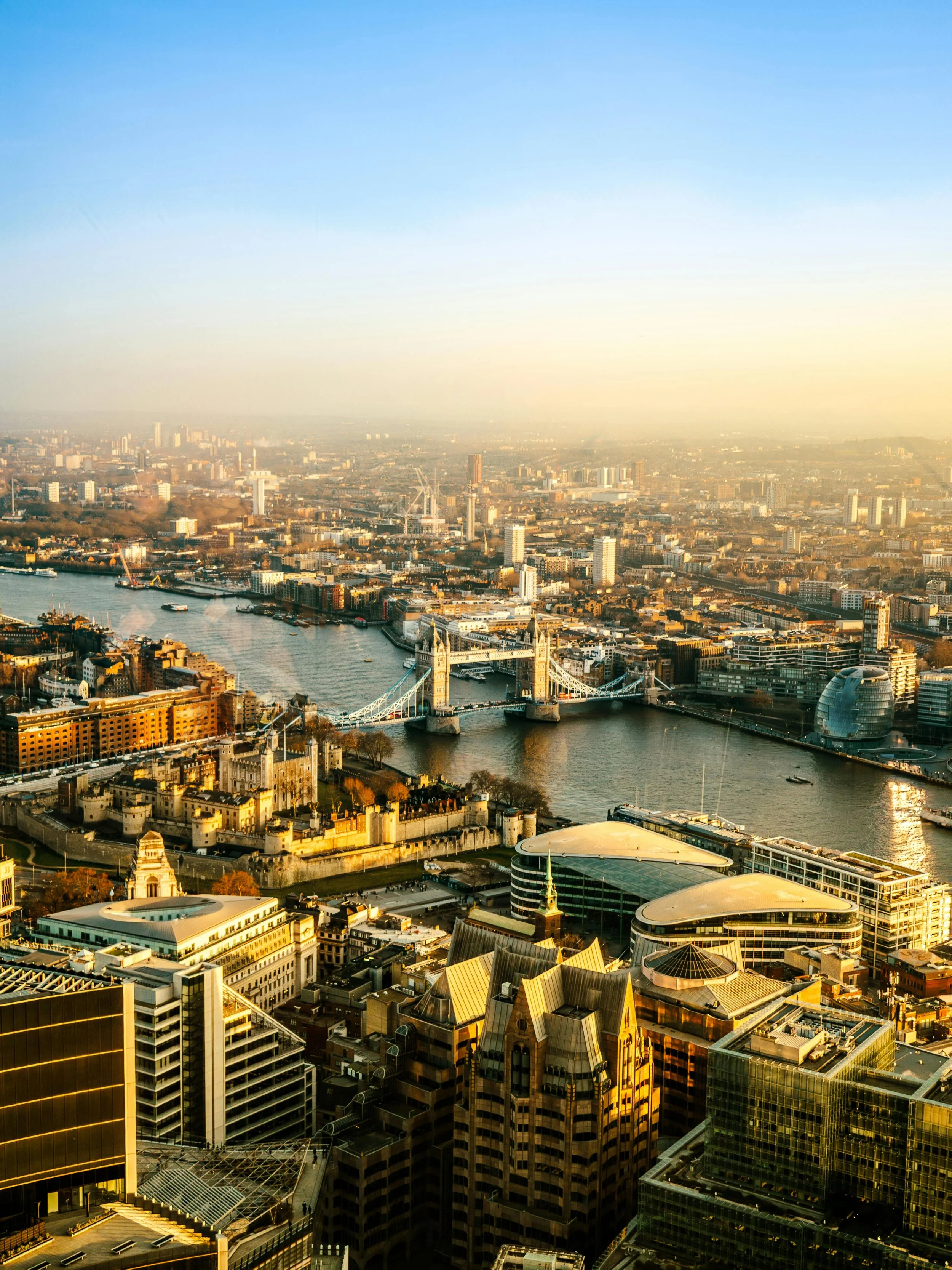 Aerial view of London cityscape with Tower Bridge in the foreground, River Thames flowing beneath, modern skyscrapers, and historic buildings on a clear day.