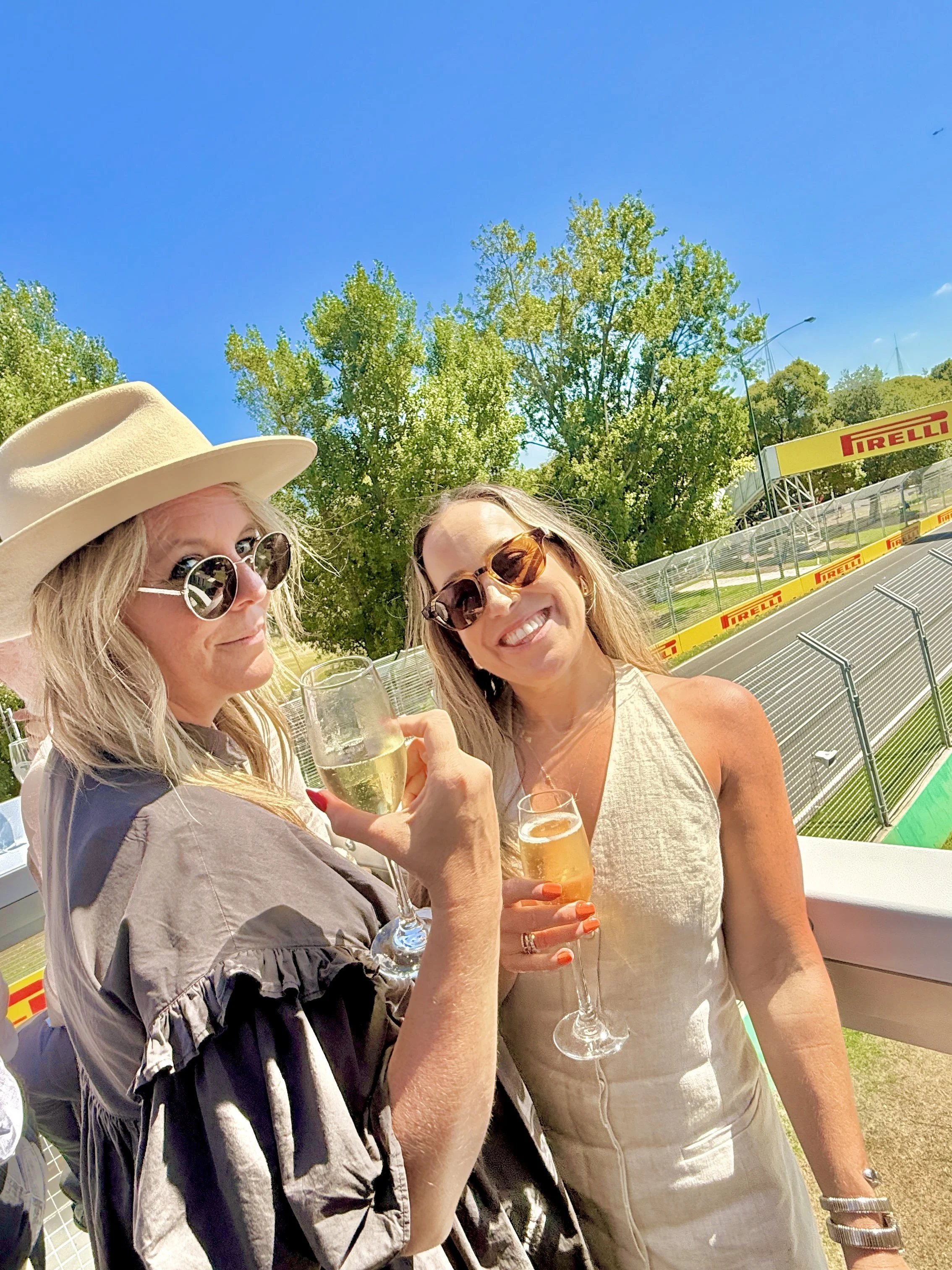 Two women at a Formula 1 race holding glasses of champagne, smiling trackside in hospitality at the Australian Grand Prix, Pirelli banners in the background.