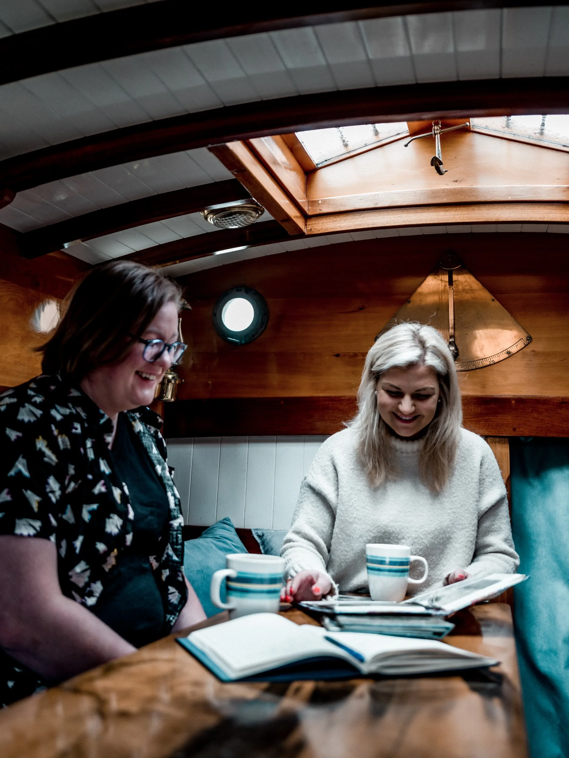 Two women sitting at a wooden table inside a cozy room with wooden interior walls, looking at a magazine and smiling, with coffee mugs and notebooks on the table.