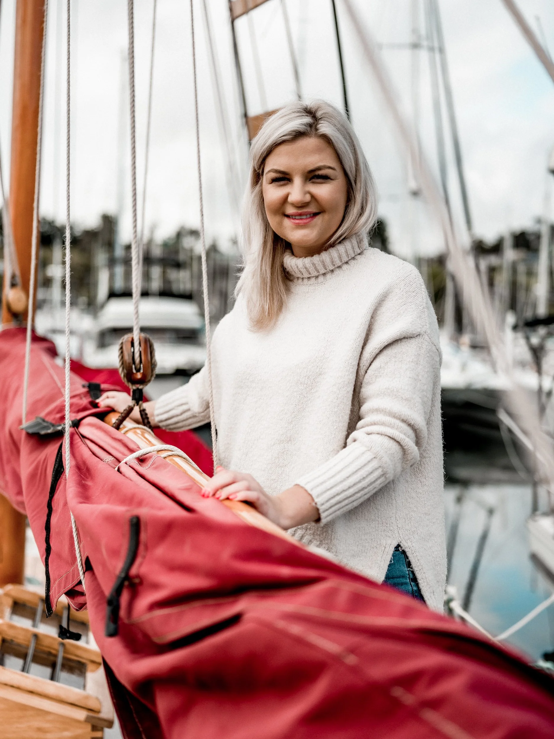 A woman with blonde hair and a beige turtleneck sweater standing on a boat, holding a red sailing bag.