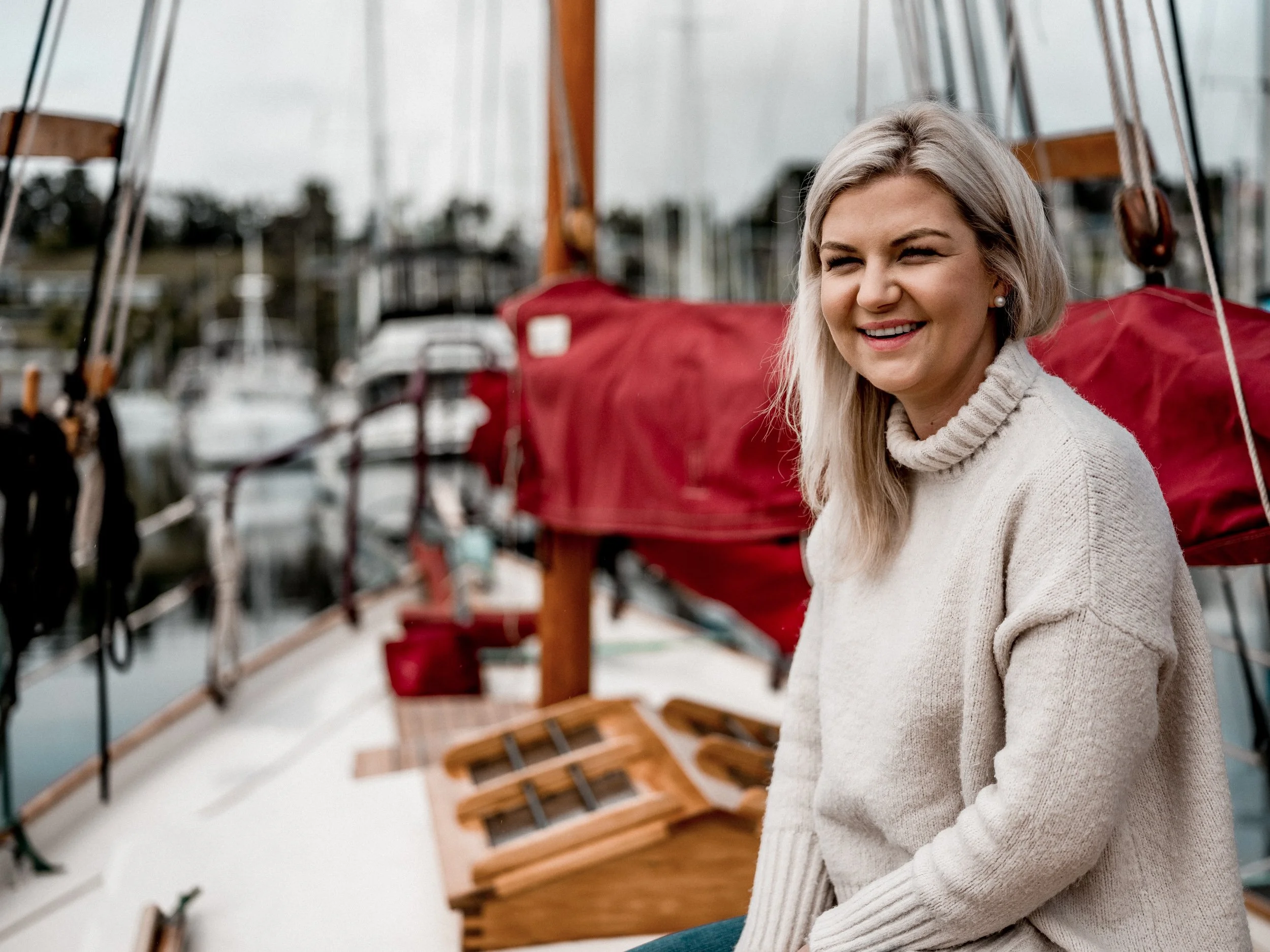 A woman with blonde hair smiling outdoors near boats and marina with blurred background.