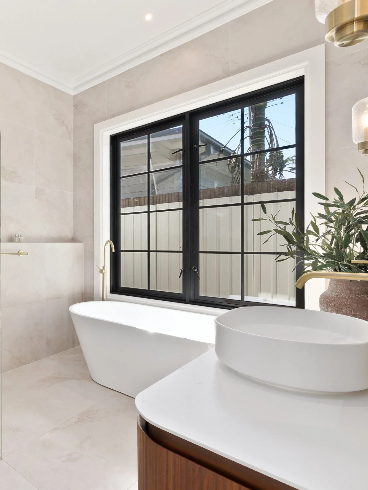 Modern bathroom with a freestanding bathtub next to a large window with black framing, beige tiled walls, a potted plant, and a vessel sink with a wood cabinet.