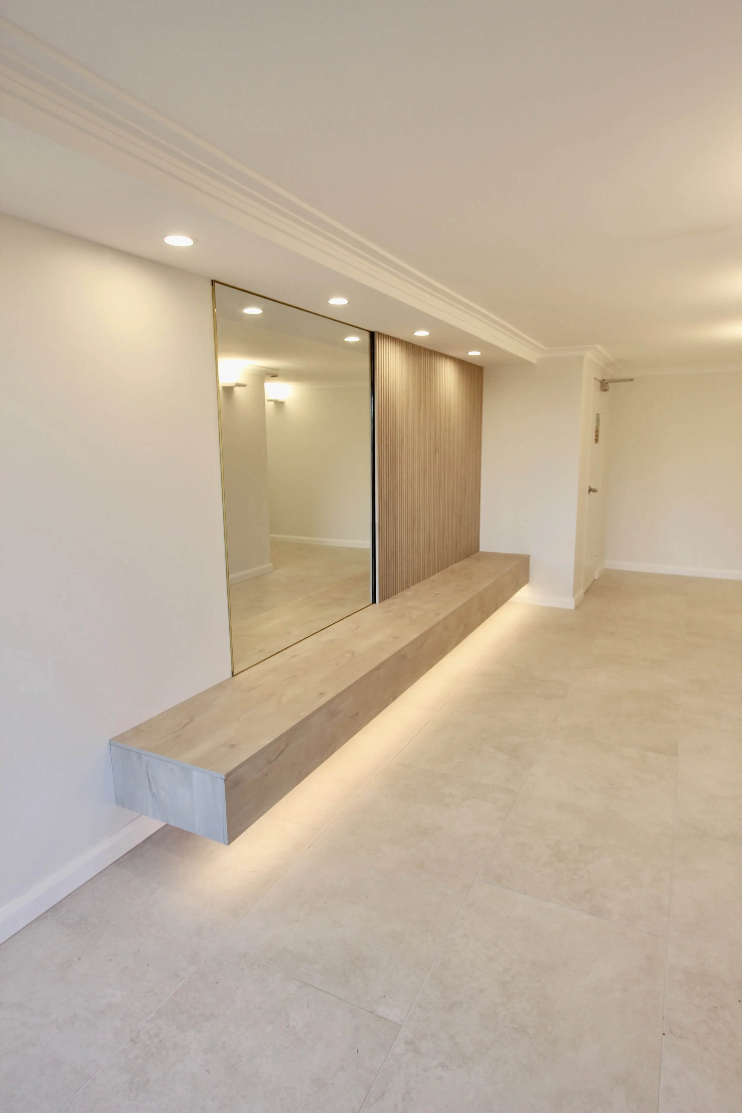 Welcoming foyer, neutral-colored tile flooring, wall-mounted wooden bench seat, a large brass-framed mirror, Screenwood acoustic wall feature. Recessed ceiling lights illuminate the space, creating a modern, clean aesthetic.