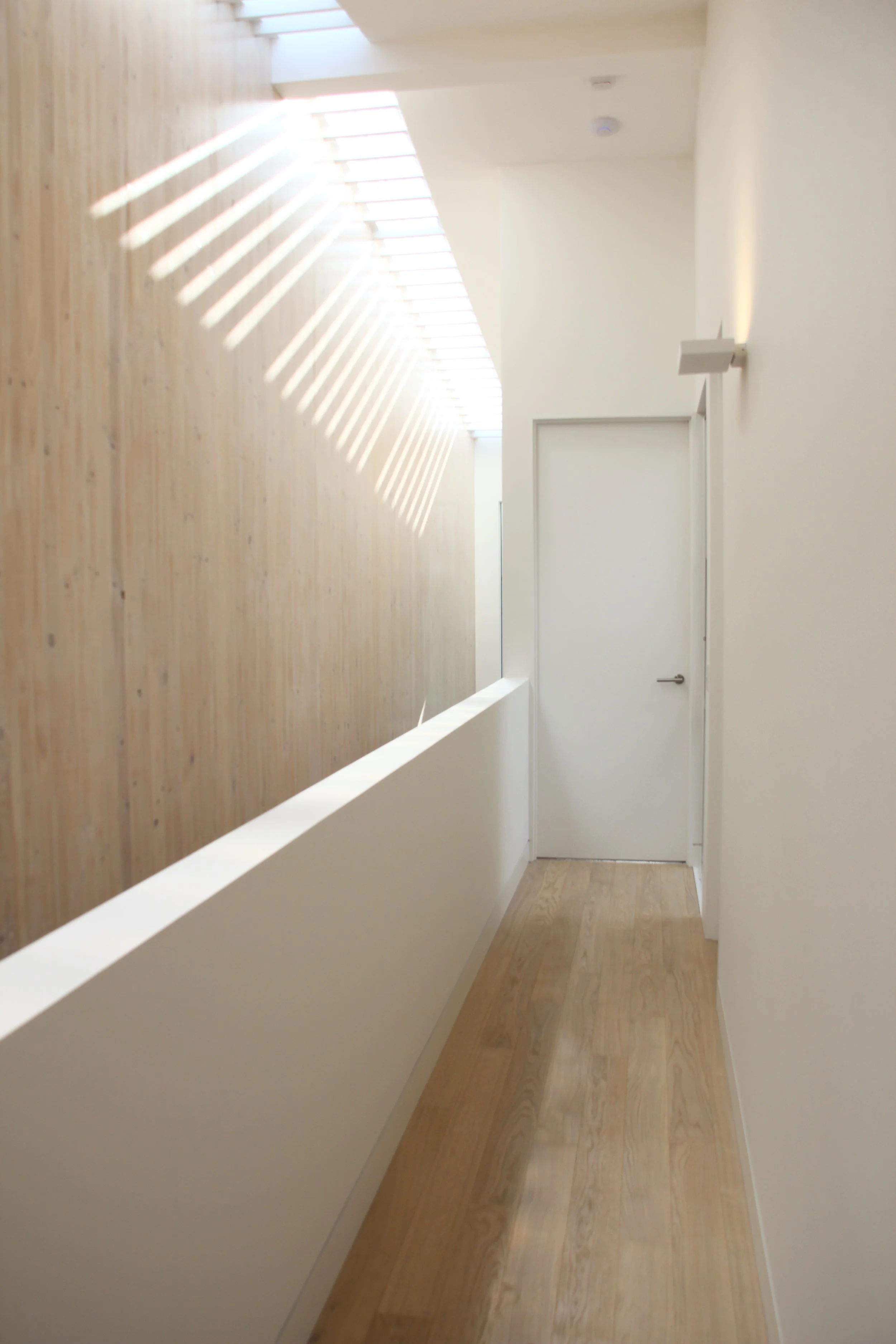 A hallway with beige wooden flooring, white walls, and a wooden accent wall on the left. Sunlight streams through a skylight, creating striped light patterns on the wall. A closed white door is at the end of the hallway.