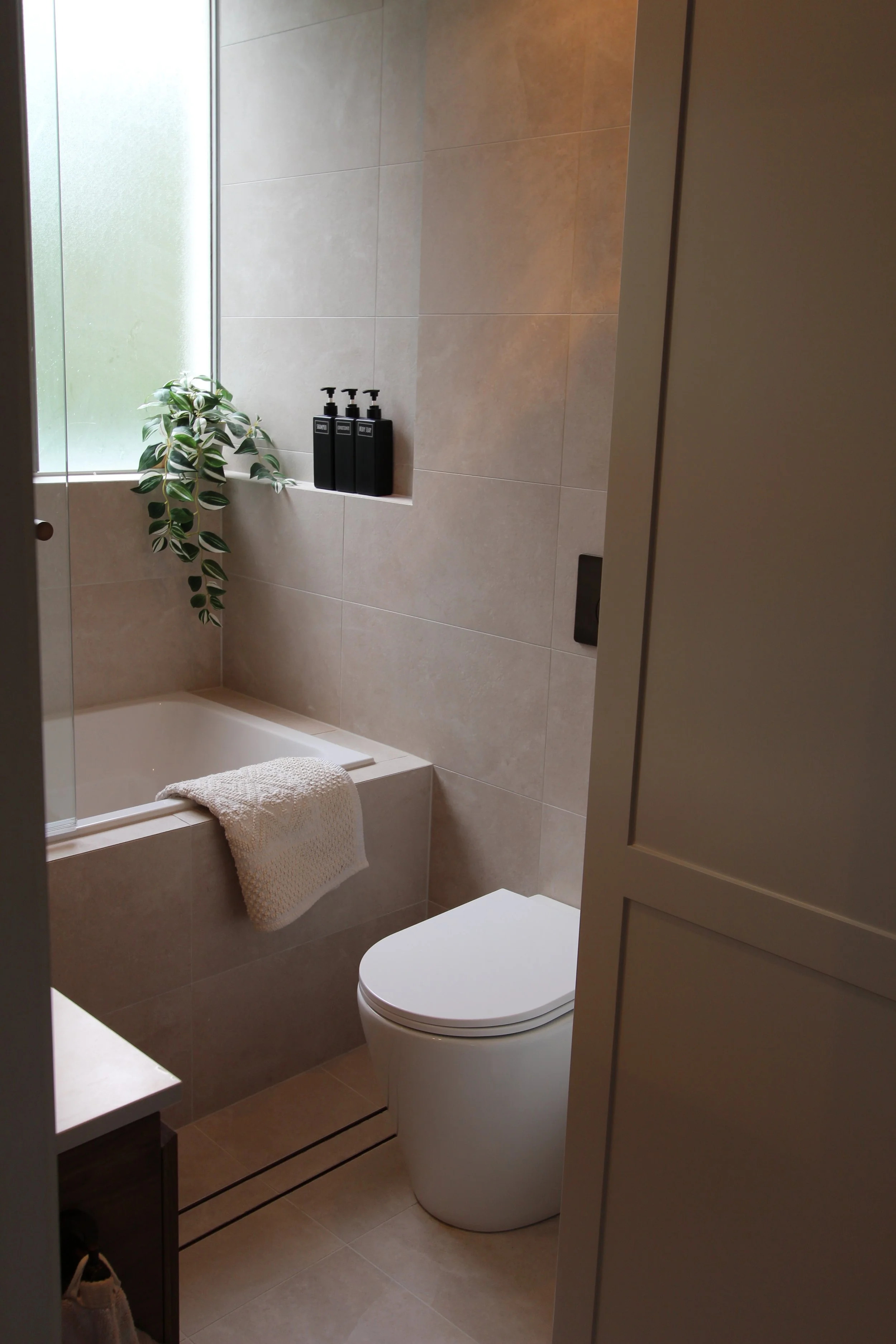 A modern bathroom with beige tiled walls, a small bathtub with a beige towel draped over its edge, a white toilet, black soap dispensers on the window ledge, a green leafy plant, and frosted glass window allowing natural light.