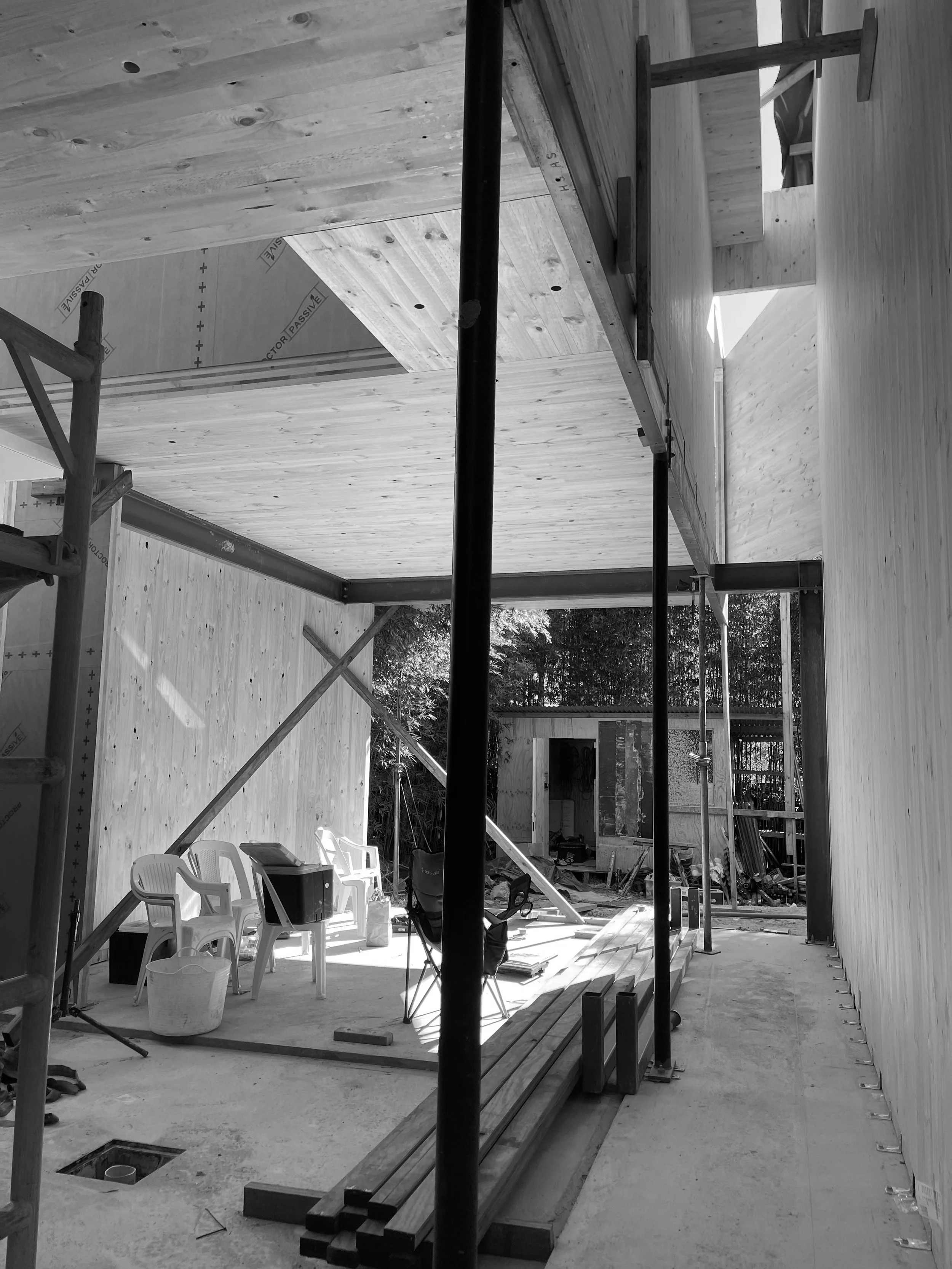 Construction site with wooden walls and ceiling, scaffolding, chairs, construction materials, and trees outside.