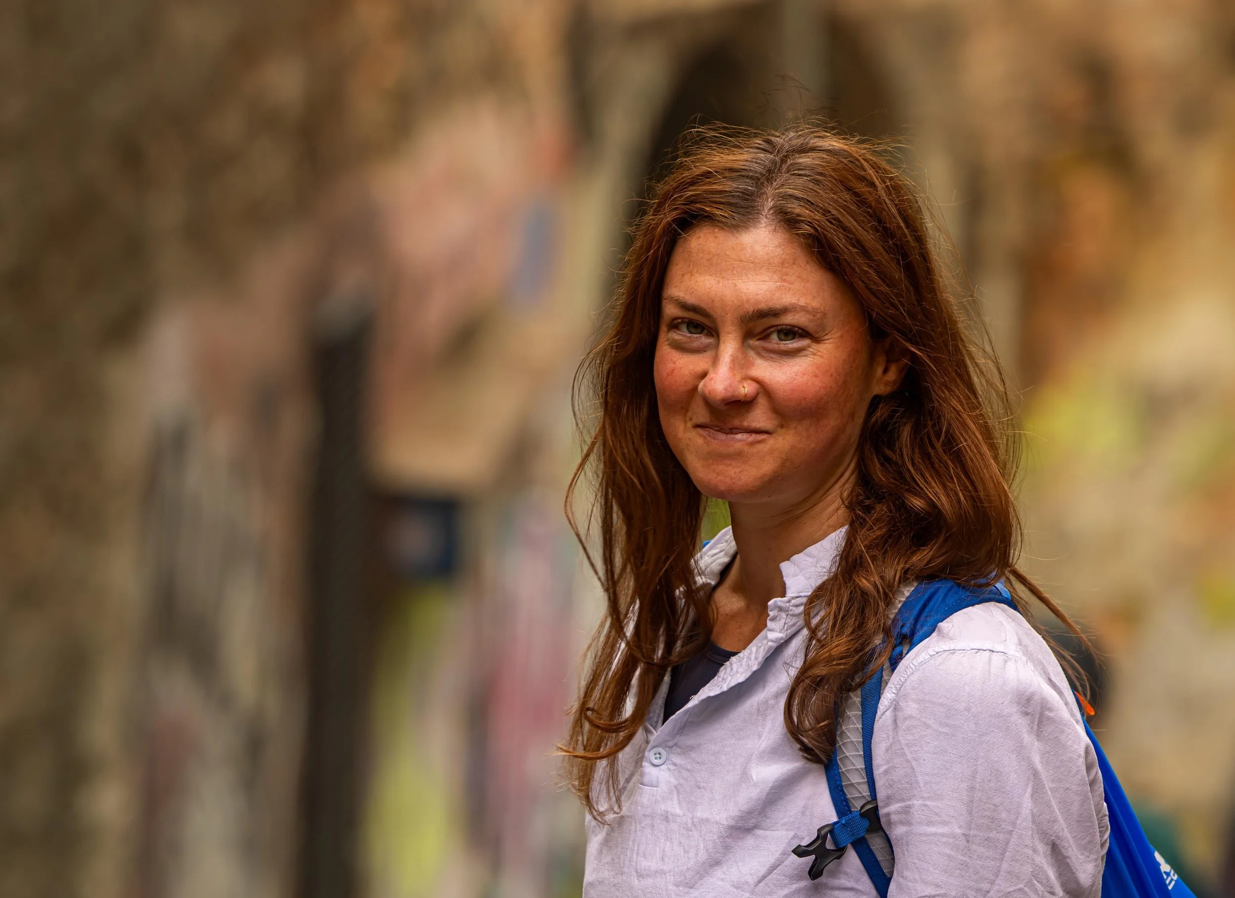 A woman with long, wavy red hair, wearing a white denim shirt, carrying a blue backpack, standing outdoors with trees in the background.