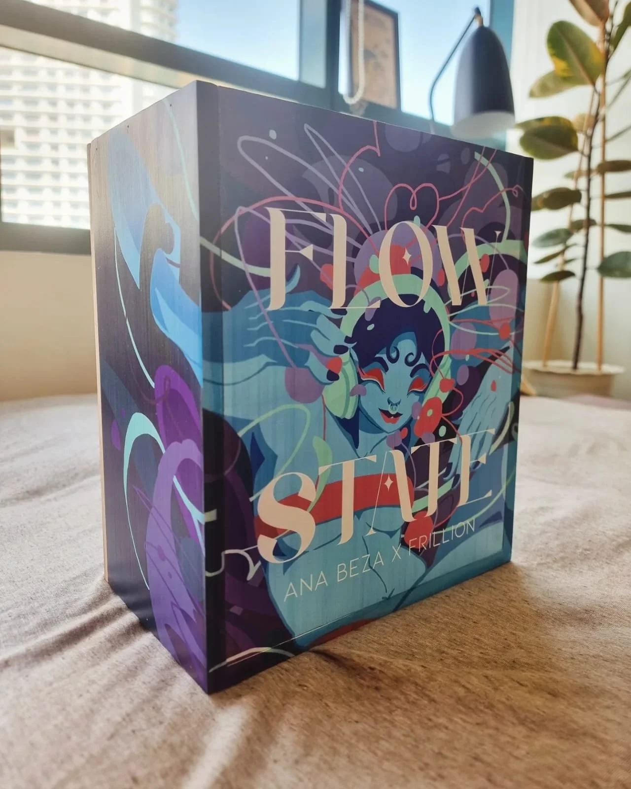 A colorful board game box with the title "Flow Stuk" on the front, featuring an illustration of a smiling woman surrounded by abstract, flowing shapes and lines. The box is placed on a beige surface near a window and a potted plant.