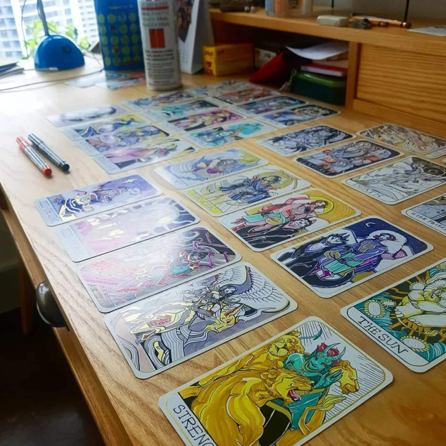 Colorful tarot cards laid out on a wooden table with pens nearby, in a room with a window and various objects on a bookshelf.