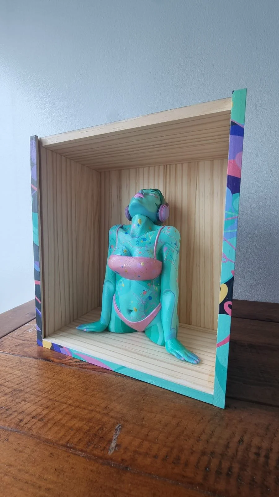 A colorful, abstract sculpture of a woman with teal skin, closed eyes, and headphones, posed as if enjoying music, encased in a wooden display box with a decorative, multicolored frame.