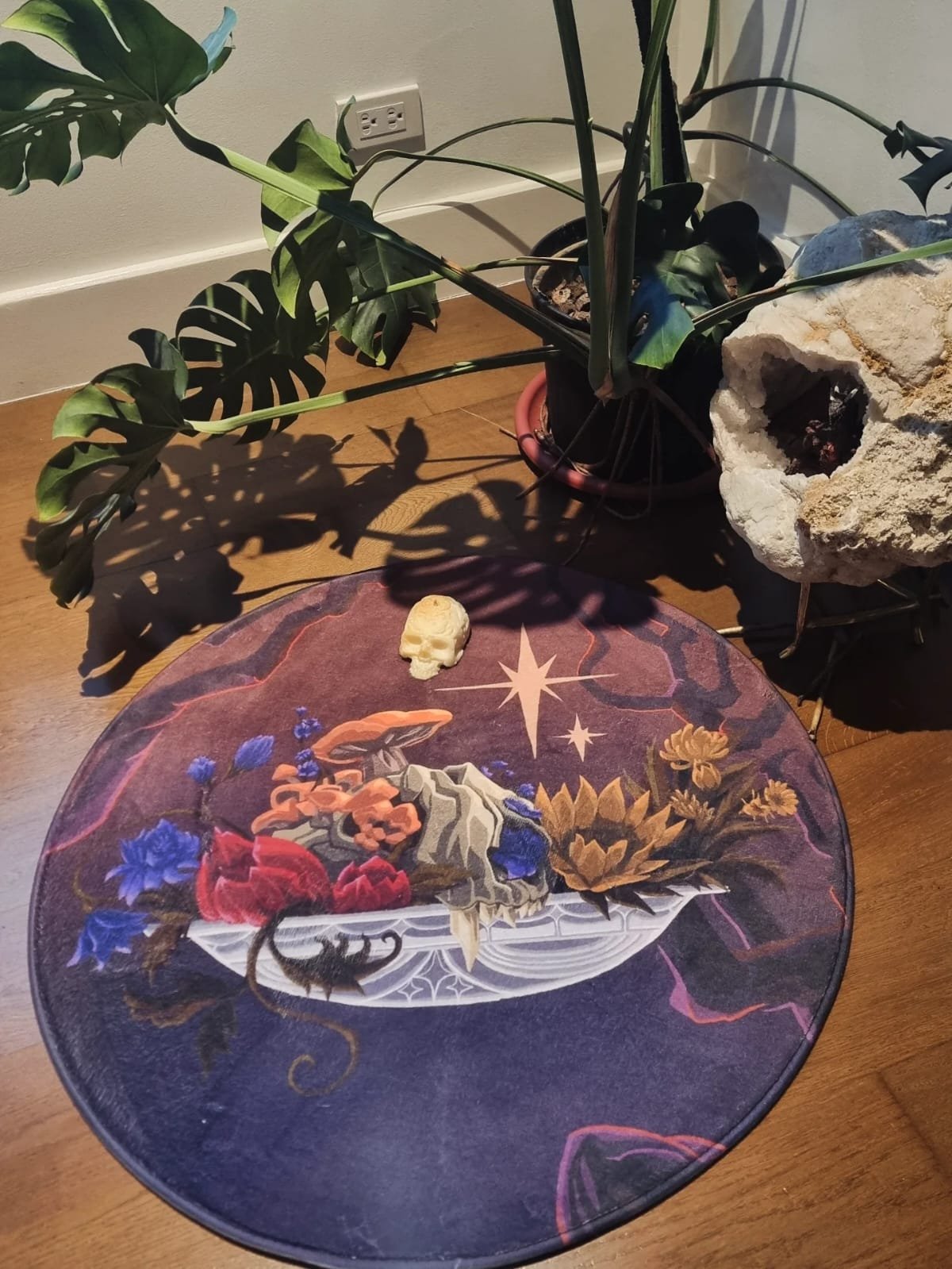 A wooden table with a colorful fantasy-themed round cloth featuring a skull with flowers, stars, and a wand design. On top of the table, there is a small beige skull figurine. Behind the table, there are potted plants, including a monstera, positioned near an electrical outlet on a beige wall.
