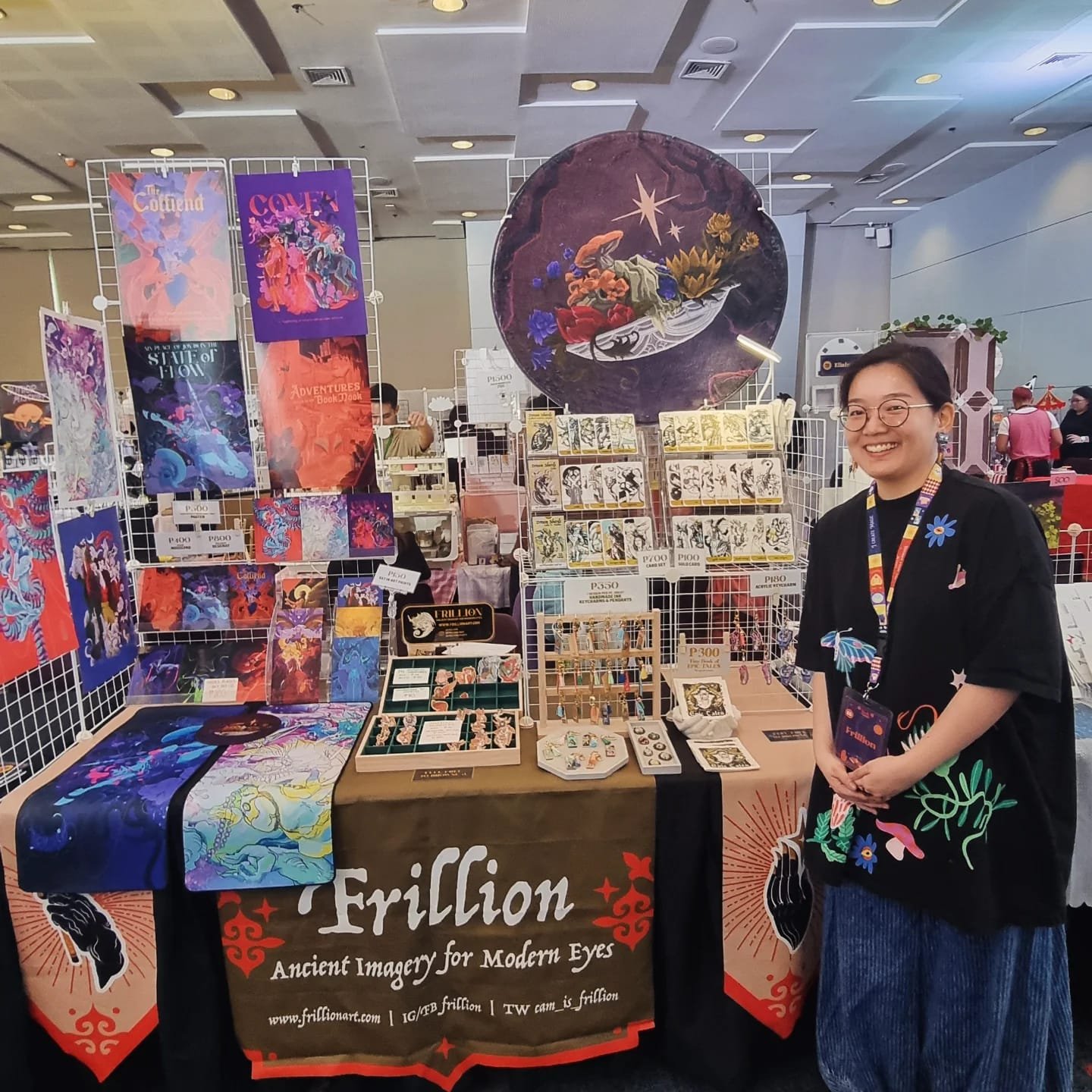 A woman with glasses and a black shirt standing in front of a booth at an art fair, with colorful prints and jewelry displayed on the table and wall behind her. The table has a sign that reads 'Friction: Ancient Imagery for Modern Eyes.'