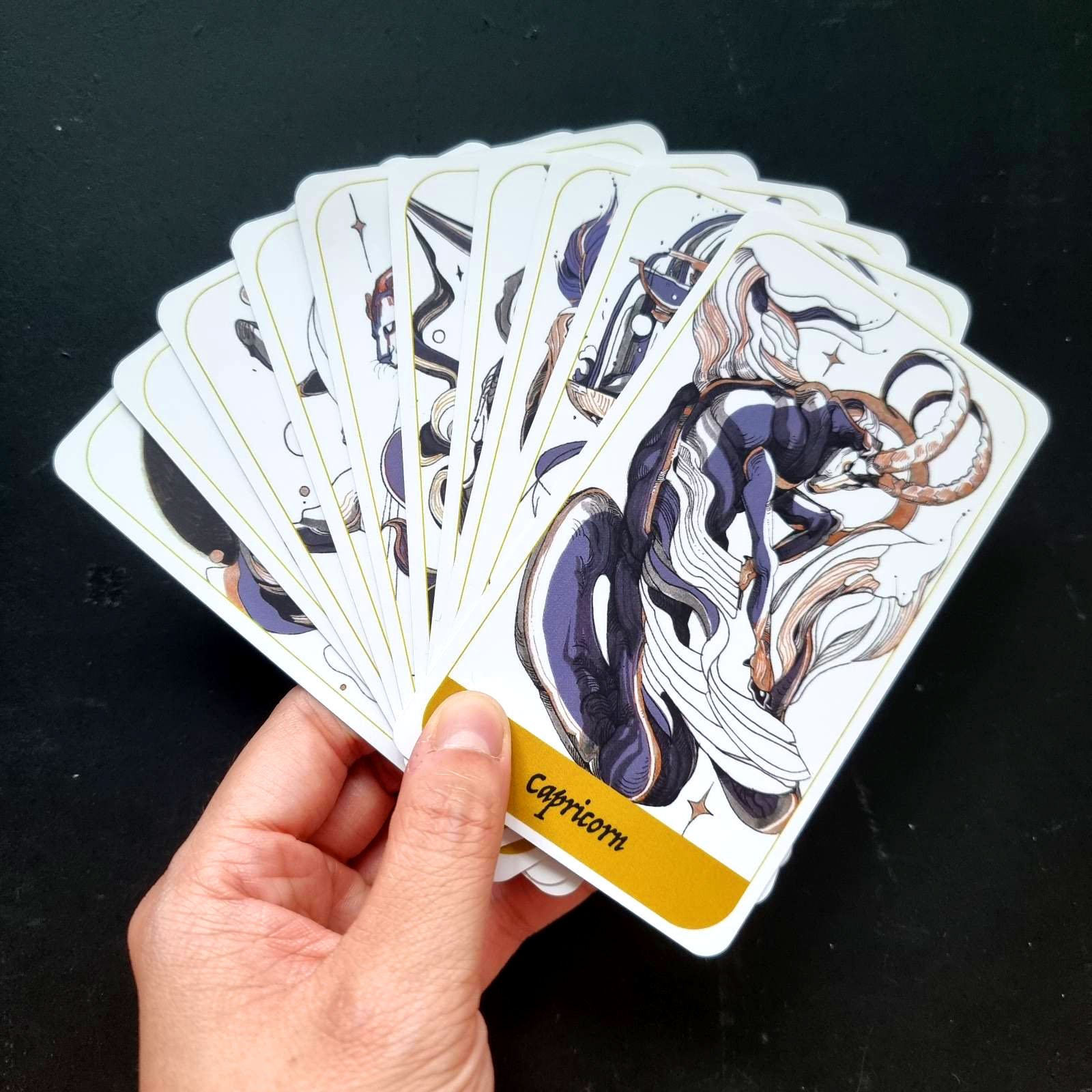 A hand holding a set of illustrated tarot cards, including the card 'Capricorn,' against a dark background.
