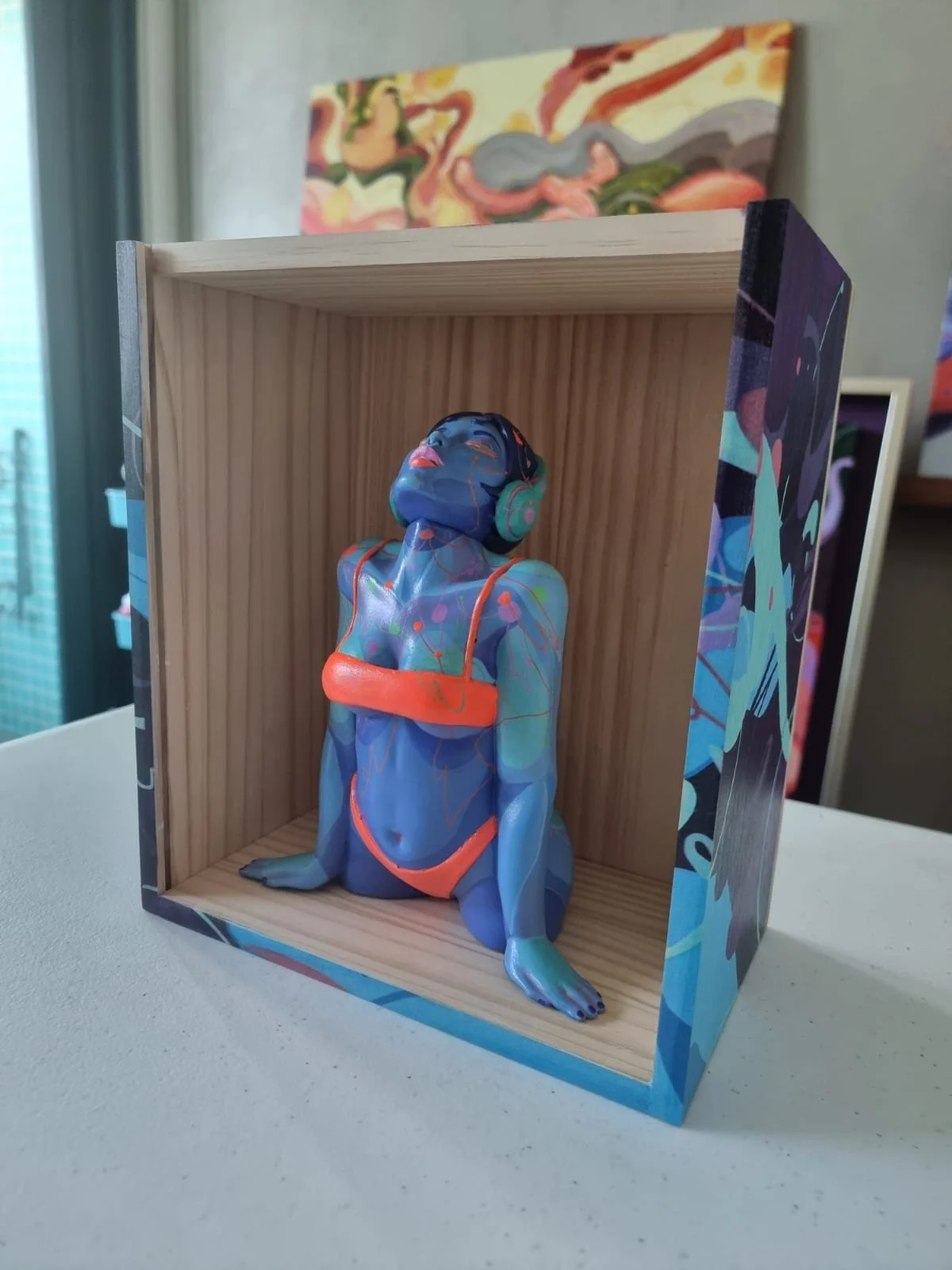 A colorful figurine of a woman with long hair, wearing a swimsuit and headphones, sitting with her legs crossed inside a wooden box, with abstract art in the background.