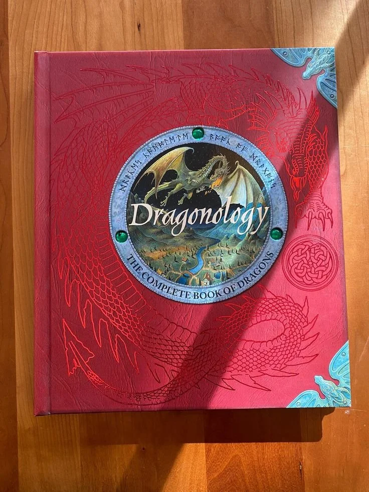 A large red hardcover book titled "Dragonology" with a circular illustration of a dragon and landscape on the cover, surrounded by red dragon illustrations and decorative elements.