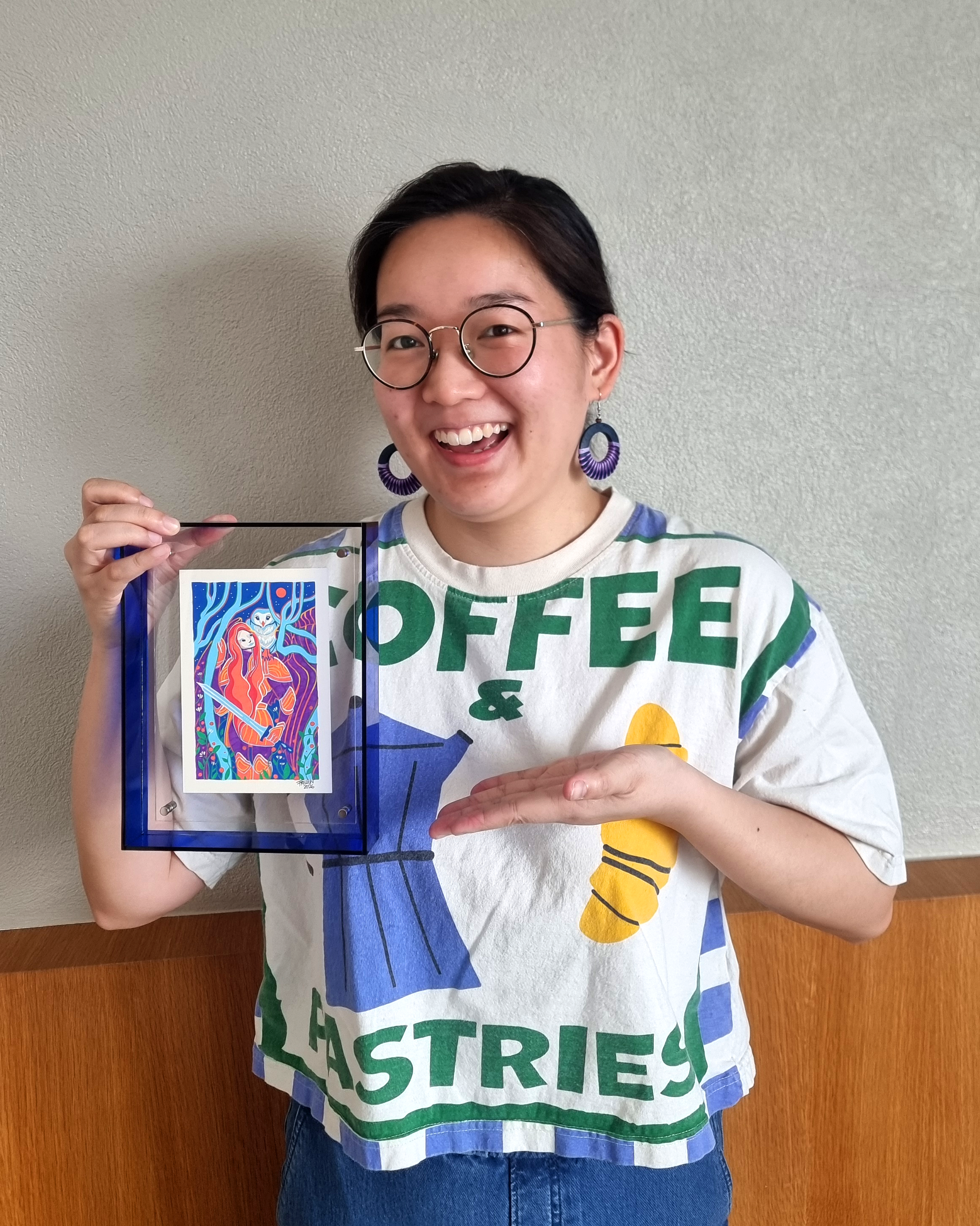 A person with short dark hair, glasses, and large purple earrings is smiling and holding a colorful artwork framed in glass. They are wearing a white T-shirt with green and yellow text and a blue and green design, standing against a plain wall and a wooden surface.