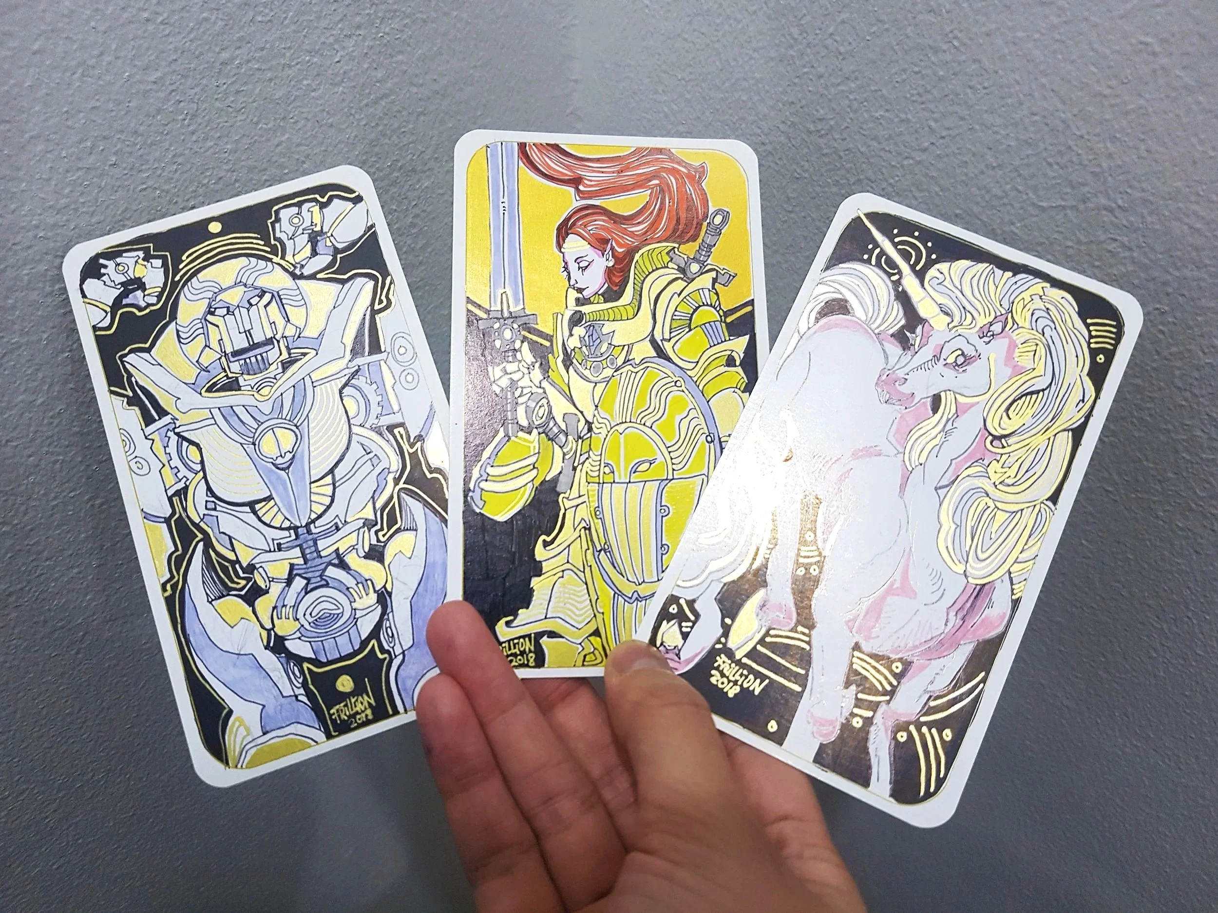 Close-up of three trading cards depicting characters with elaborate, futuristic armor and art styles, held by a person's hand against a gray background.