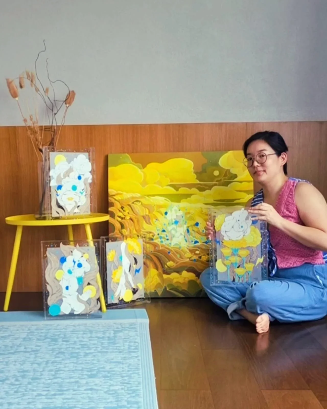 Woman sitting on the floor beside a yellow table displaying several abstract paintings with yellow, blue, and white colors, a large abstract landscape painting in the background, and a vase with dried plants on the table.