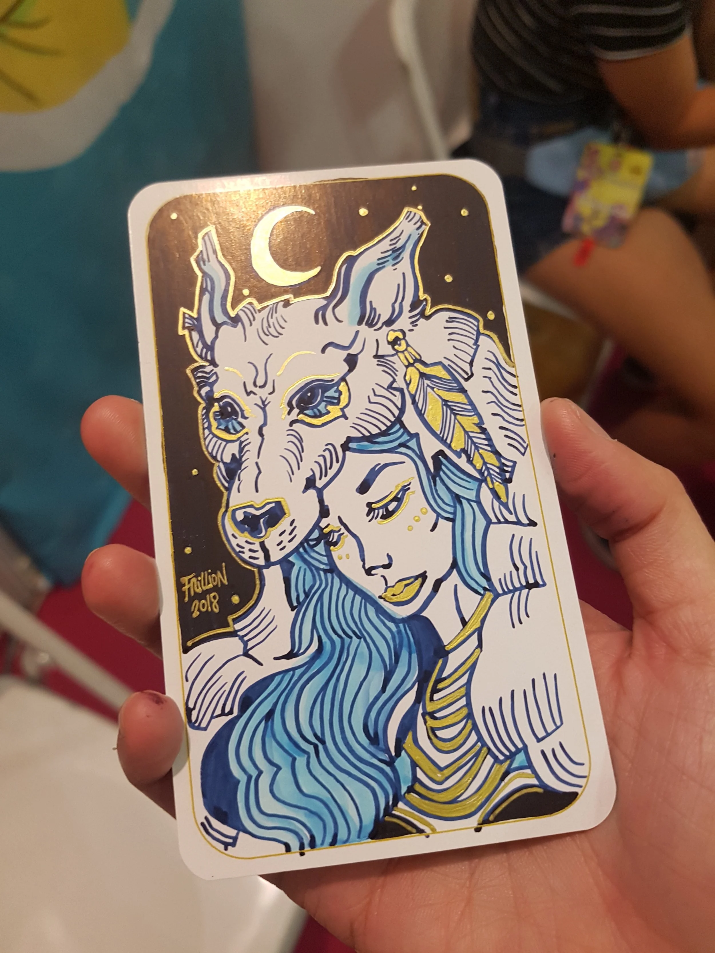 A playing card with a stylized illustration of a woman with blue hair and a dragon. The card features gold accents and is signed by 'Hillion 2018'.