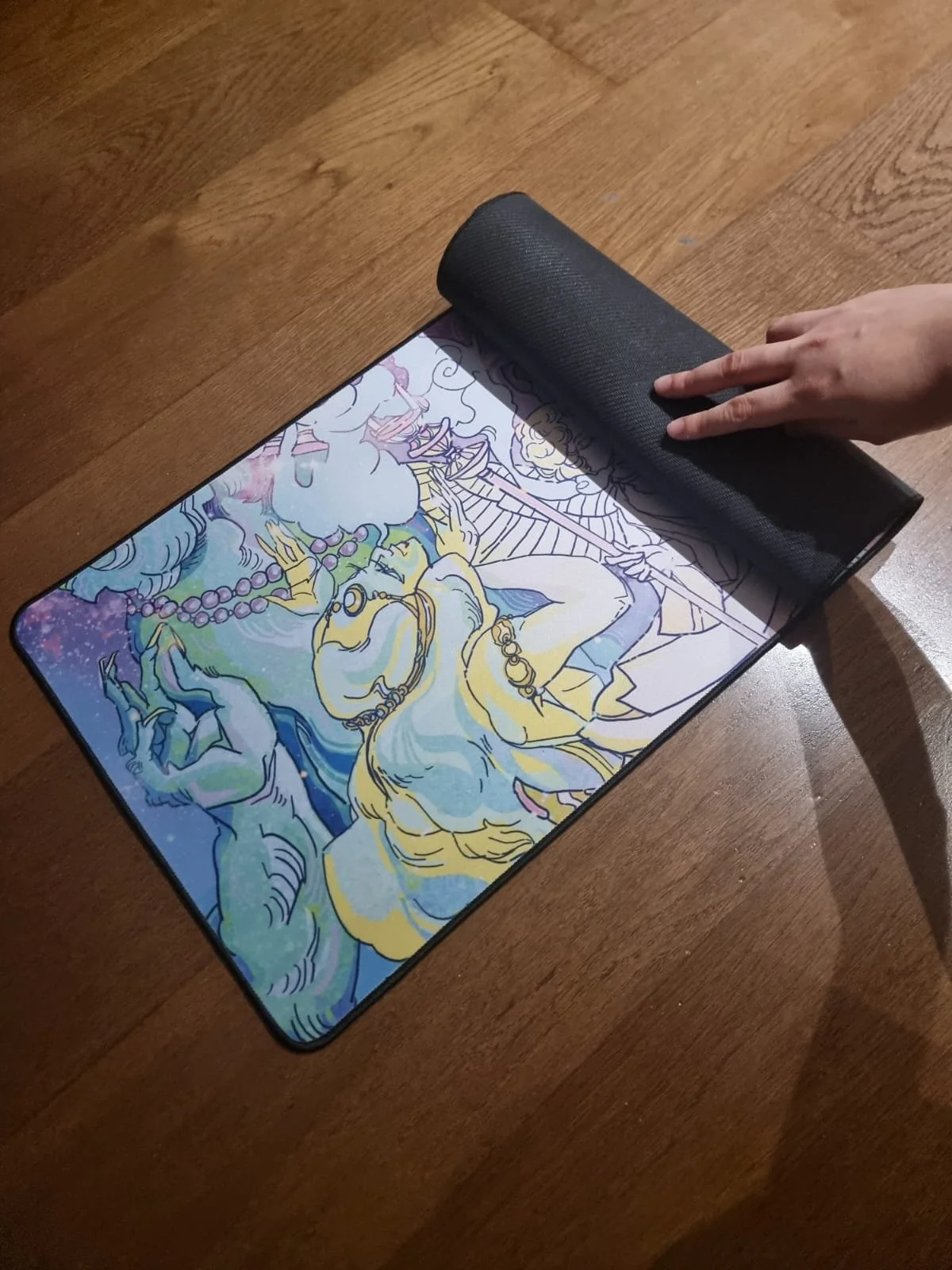 A person is lifting a black fabric cover from a tablet displaying a colorful drawing of a mythological figure, such as a mermaid, wearing jewelry and surrounded by clouds and water, on a wooden table.