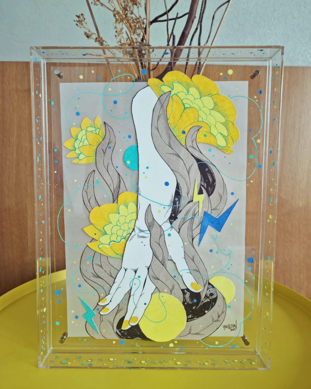 A mixed media artwork featuring a hand with painted yellow nails and black outlines, surrounded by yellow flowers, swirling blue lines, and blue and yellow lightning bolts on a transparent rectangular panel with a wooden background.