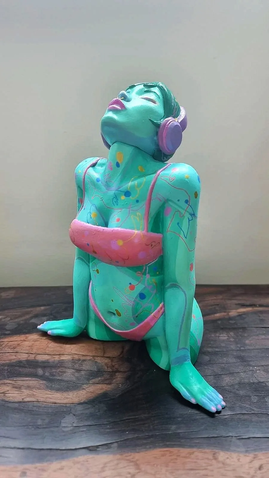 A colorful frog sculpture with a woman's face, wearing headphones, painted in pastel shades and patterns, sitting on a wooden surface.