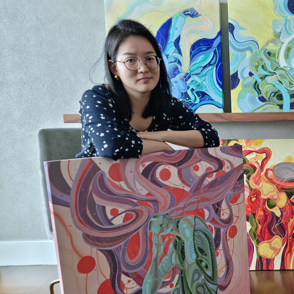 A woman with glasses and dark hair, wearing a black blouse with white stars, sitting with her arms crossed in front of abstract colorful paintings featuring swirling patterns and vibrant colors.