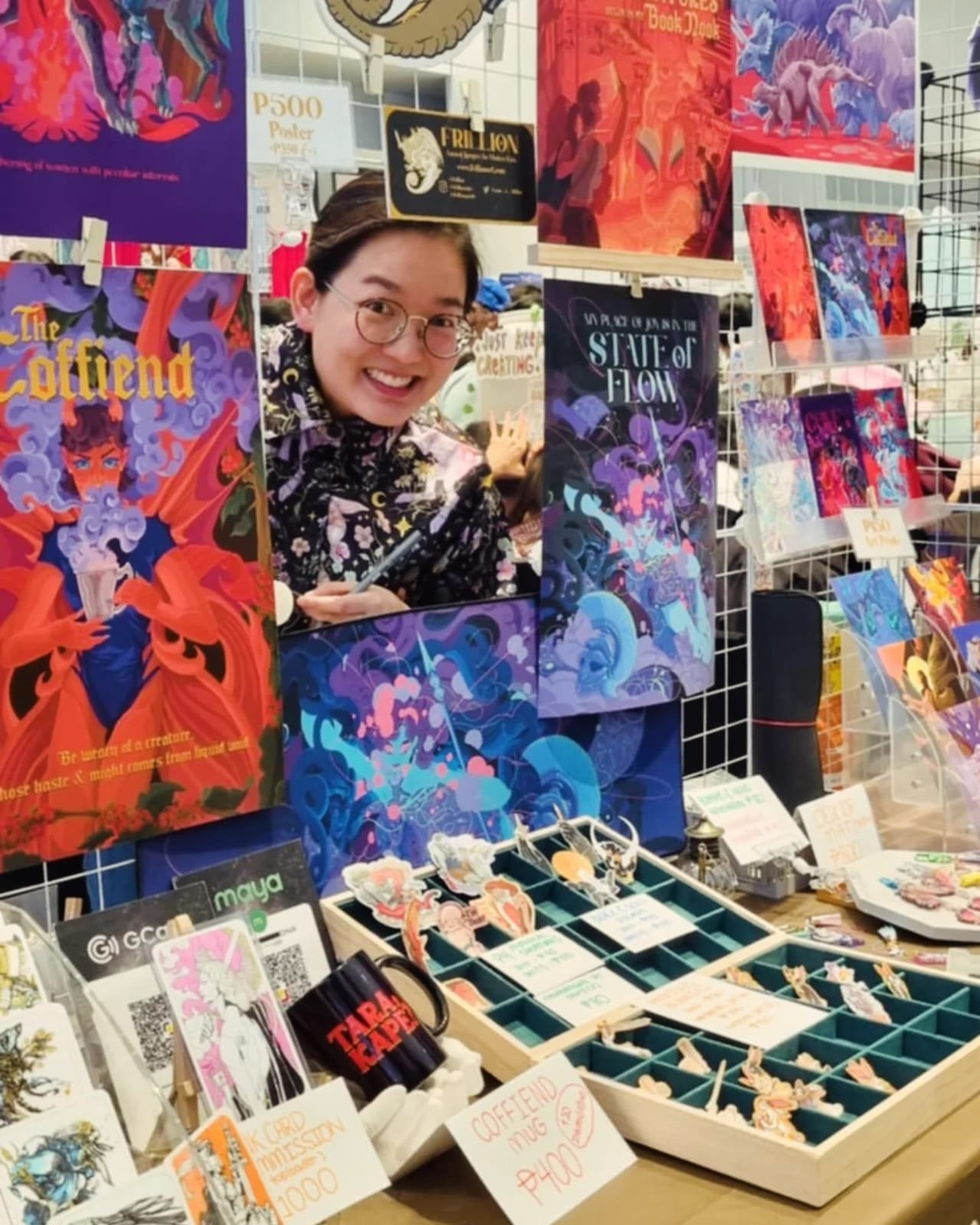 A woman with glasses smiling at a vibrant art and merchandise stall at a fair or market. The stall displays various colorful posters, pins, and small art prints with fantasy and sci-fi themes, and signs indicating prices and product descriptions.