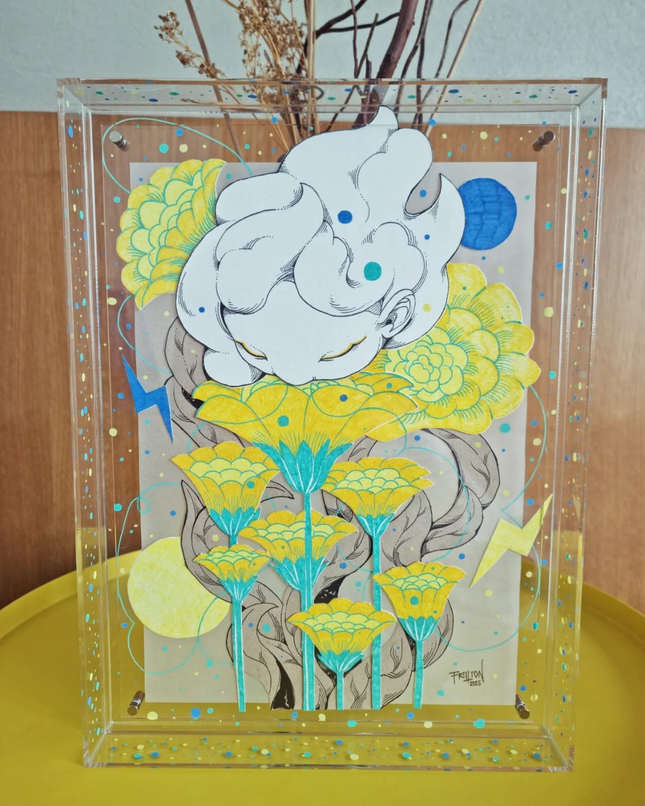 A mixed media artwork featuring a black and white line drawing of a cat's head atop yellow and blue floral elements, with abstract and celestial motifs in the background, inside a clear acrylic display case.
