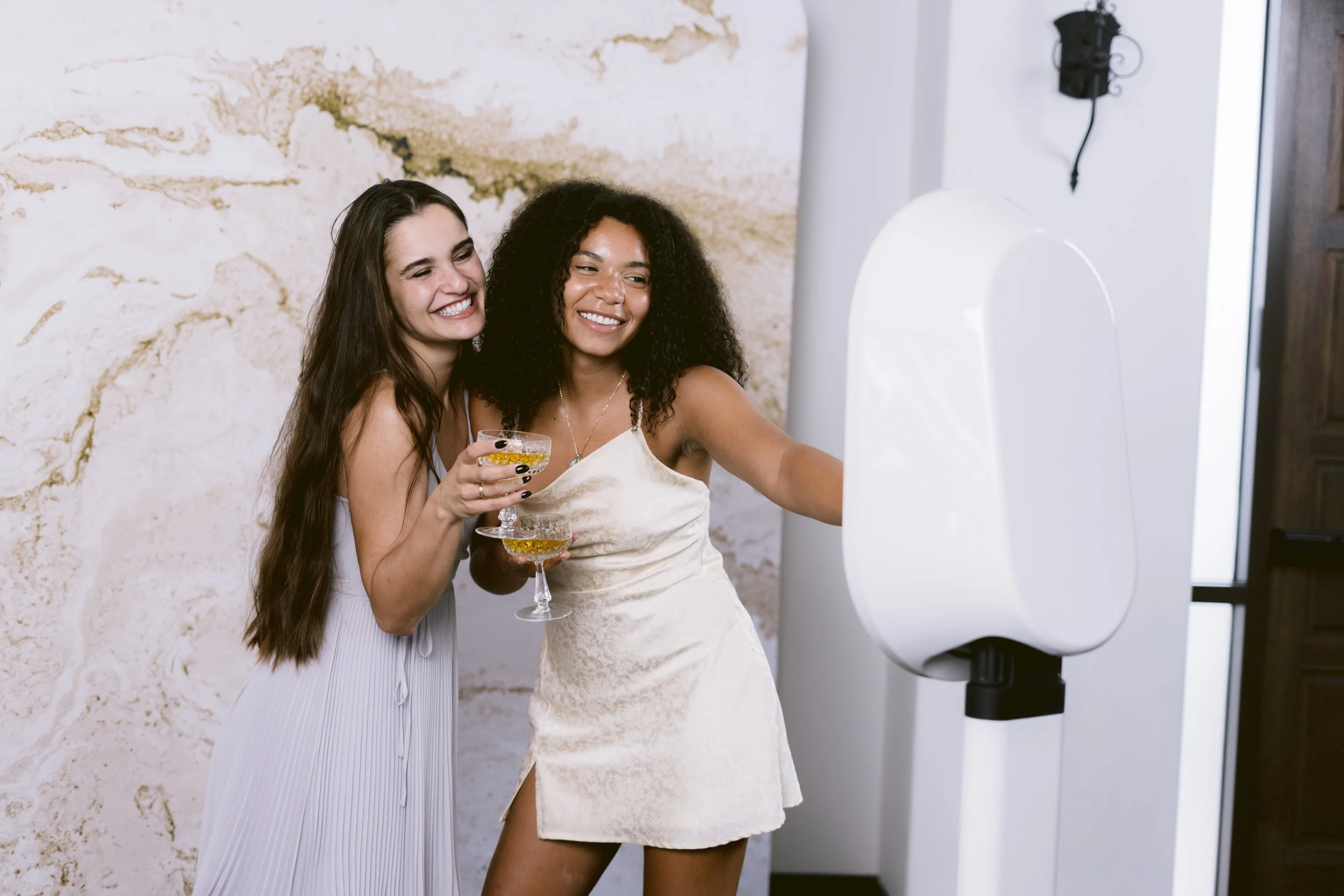 Two women smiling and taking a selfie together, holding glasses of champagne, in a modern indoor setting with a textured wall and a urinal in the background.