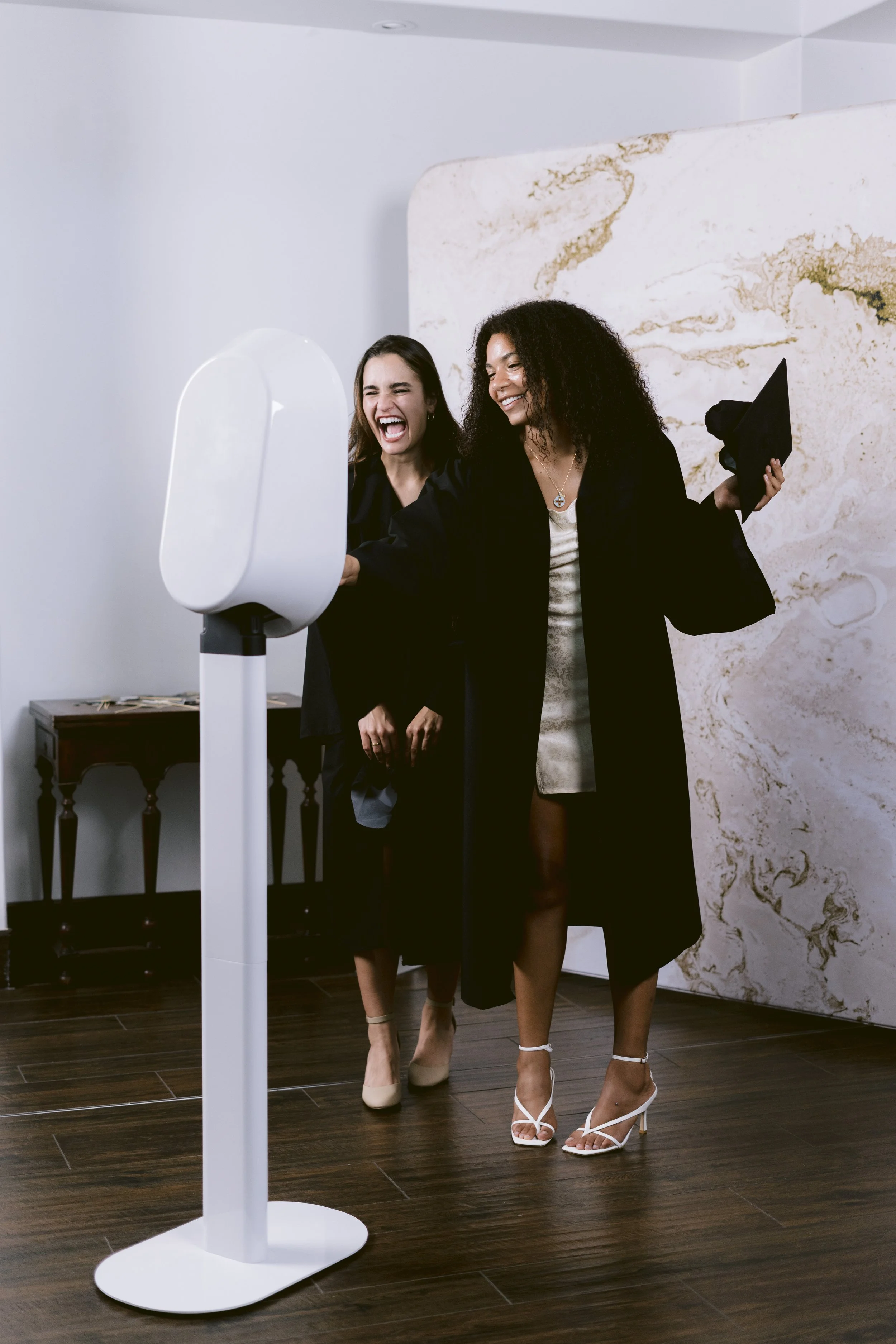 Two women are taking a selfie together with a photo booth device. They are smiling and appear to be enjoying themselves. One woman has dark curly hair and is wearing a black coat with nude heels, while the other woman has straight dark hair and is we