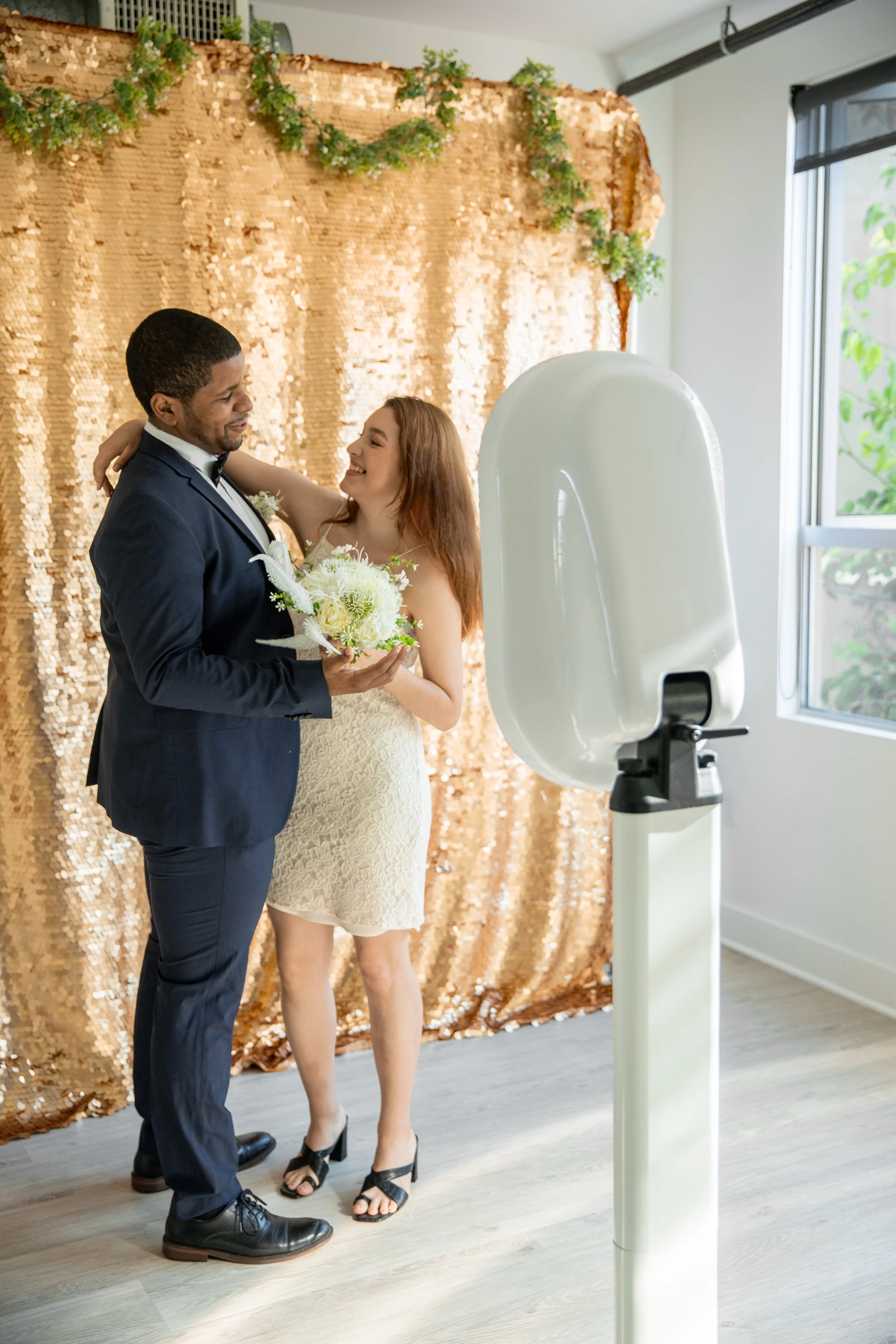 A couple dances at their wedding reception with a photo booth setup, gold sequin backdrop, and a photo booth camera in the foreground.