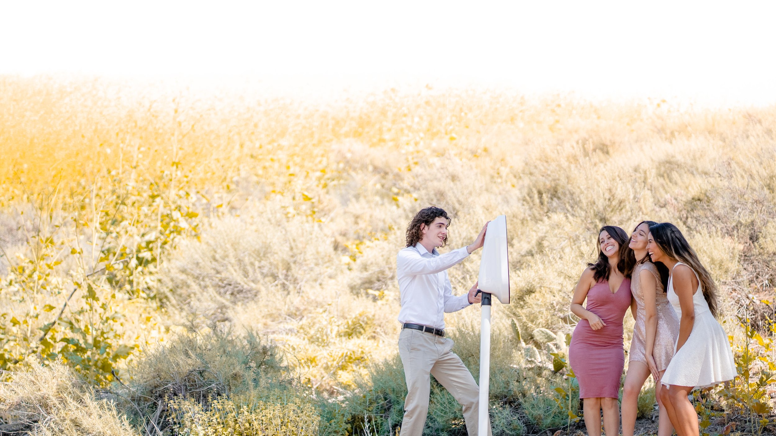 A group of women, dressed in semi-formal attire, standing outdoors in a desert-like environment with cacti and yellow foliage. They are smiling and posing for a photo taken by a young man using a camera on a tripod.