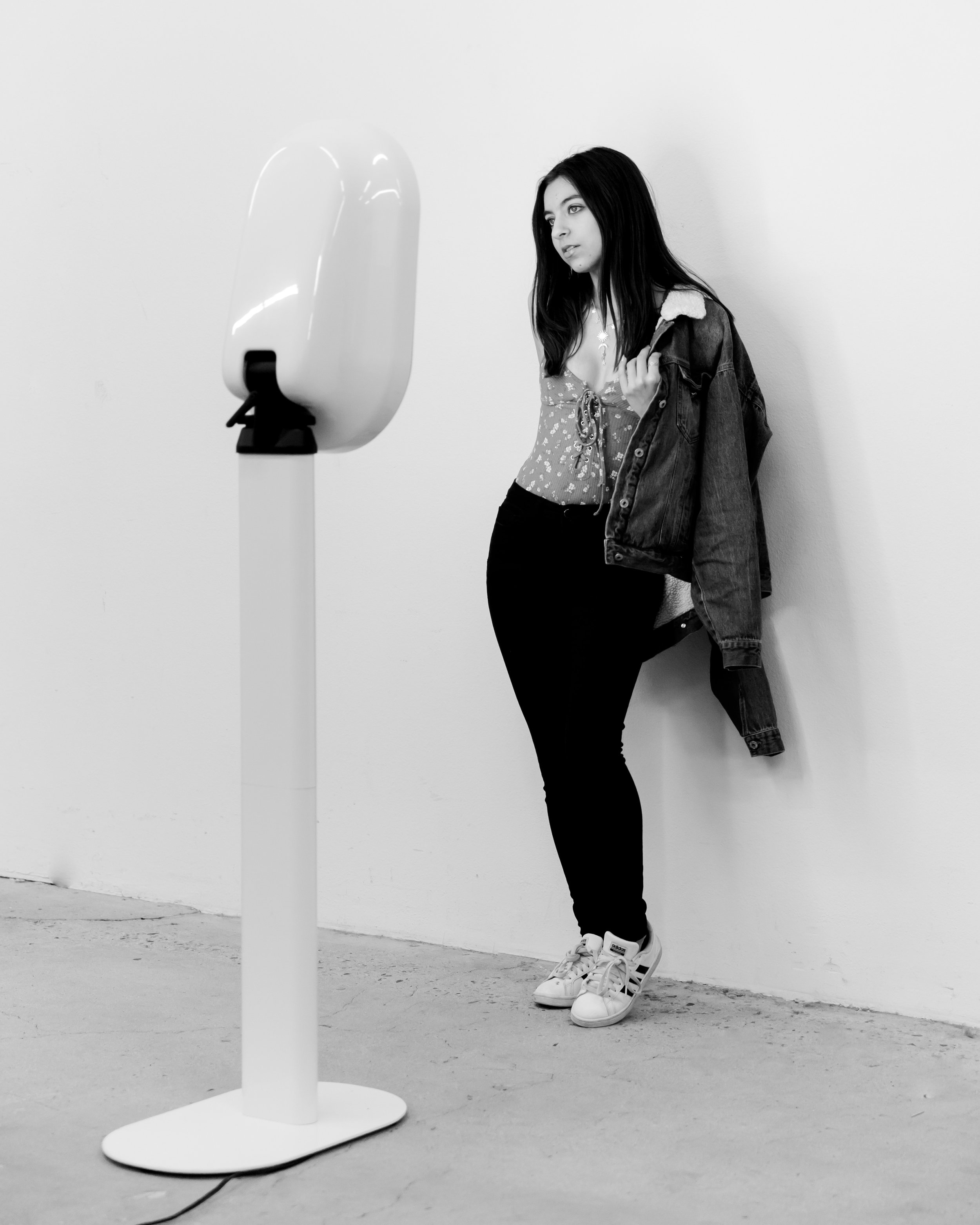 A young woman with long dark hair, wearing a patterned top, black pants, and white sneakers, stands against a plain wall, looking at a mirror with a contemplative expression. She is holding a denim jacket over her shoulder.