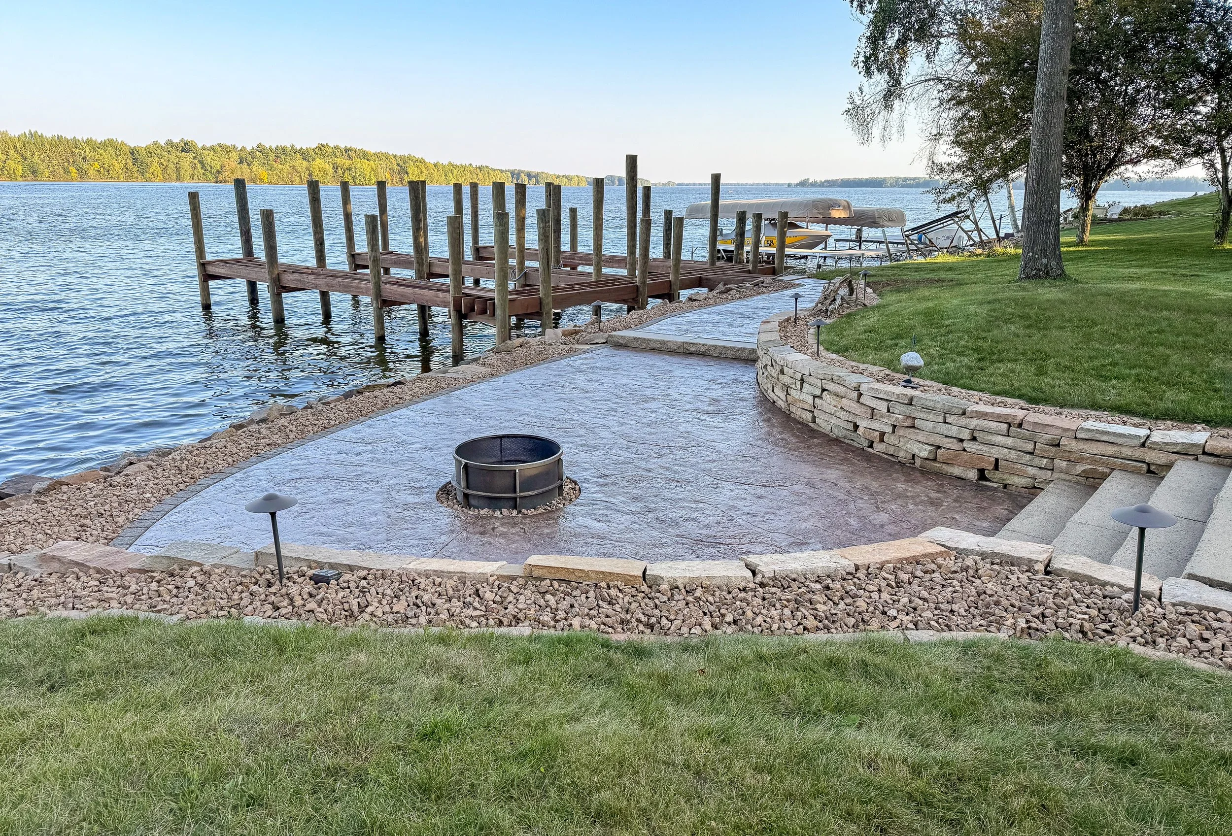 A lakeside backyard with a stone pathway, a wooden dock extending into the water, boats parked along the shoreline, a large tree, and a grassy lawn with landscape lighting.