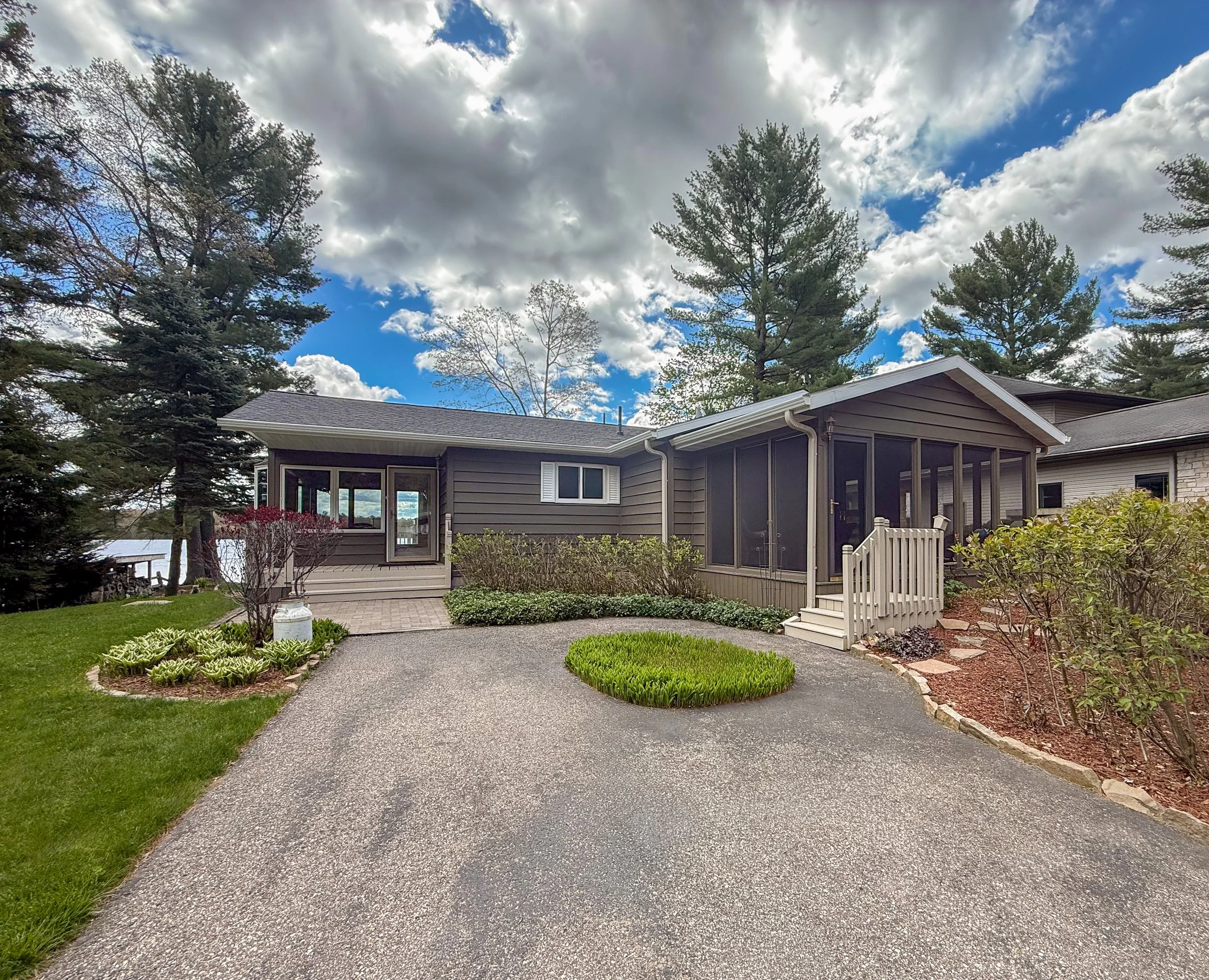 Backyard with a stone pathway, green lawn, bushes, and trees with a house in the background under a partly cloudy sky.