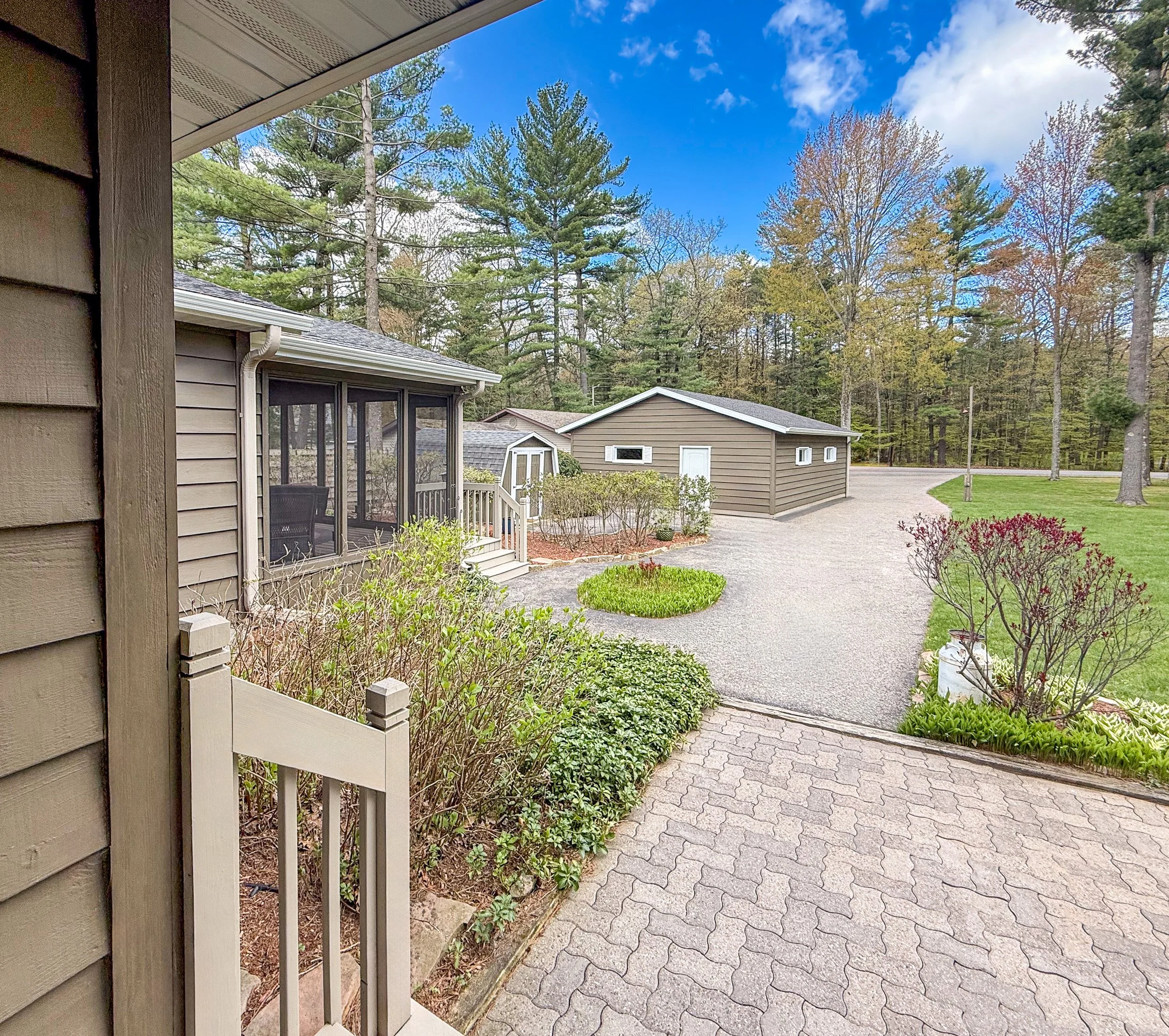 View from a house porch showing a paved walkway, grassy yard, garden, small sheds, and a wooded area with tall trees under a partly cloudy blue sky.