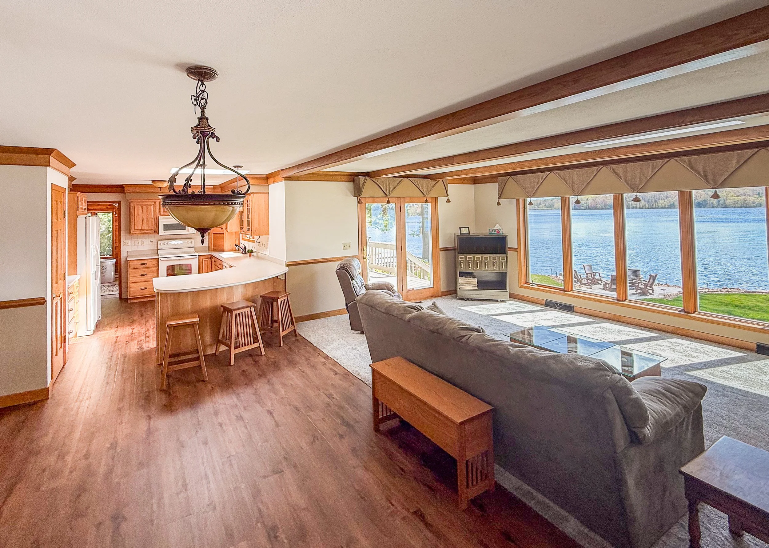 Living room with large windows overlooking a lake, wooden ceiling beams, beige walls, and wooden furnishings.