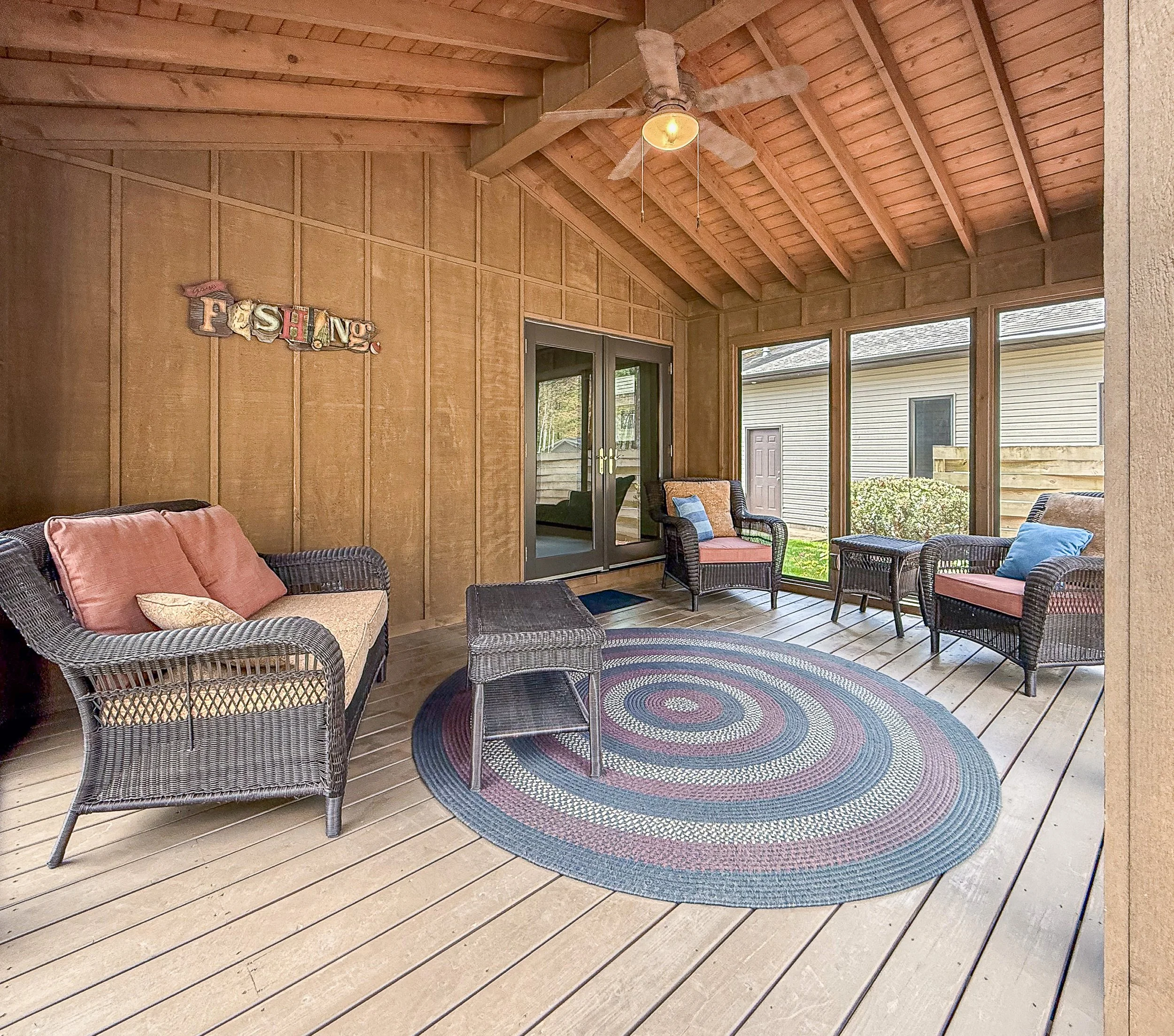 Covered outdoor patio with wicker chairs, pink cushions, a colorful braided circular rug, hardwood ceiling with a ceiling fan, large windows, and sliding glass doors.