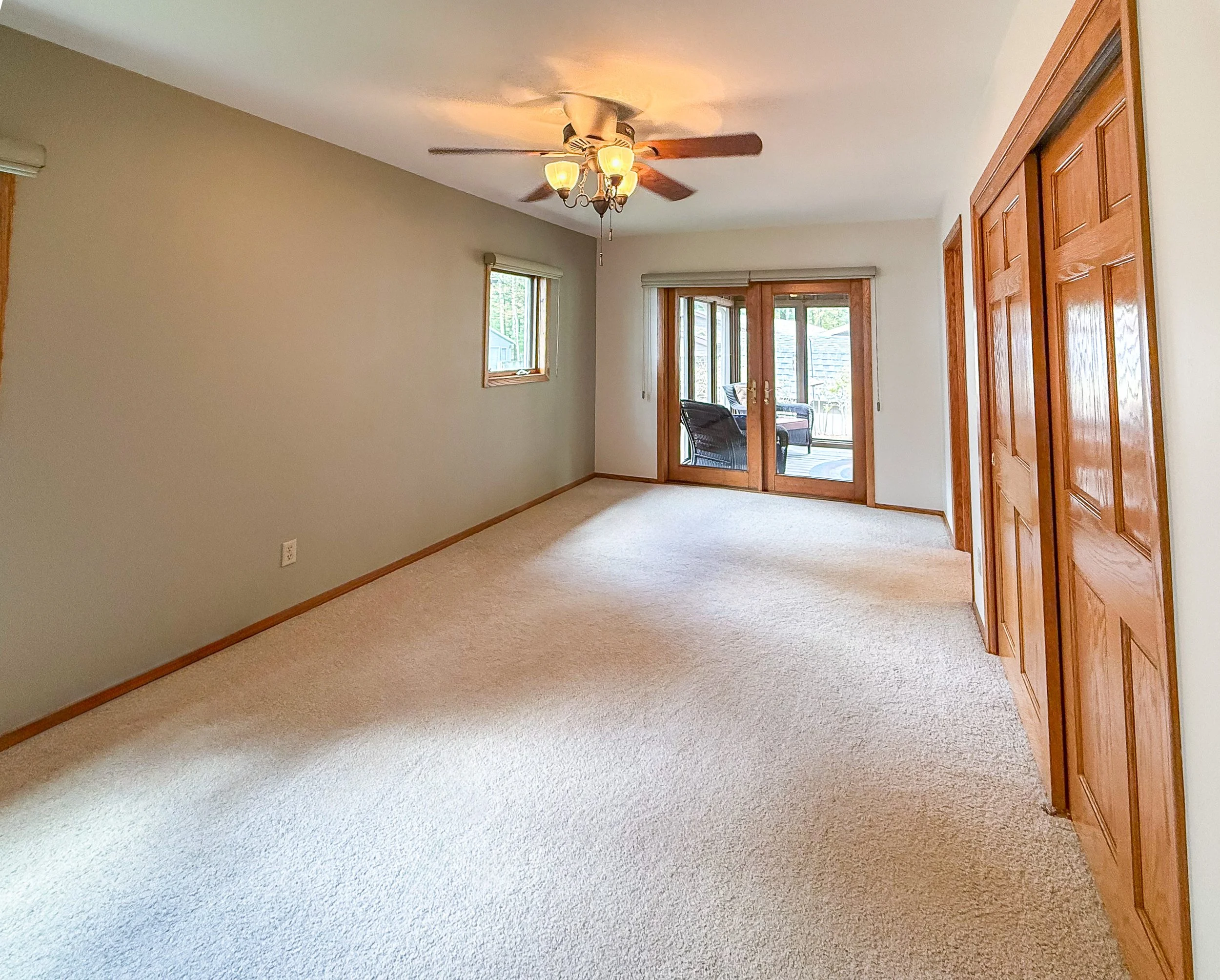 Empty living room with beige walls, beige carpet, wooden trim, windows, glass sliding door leading outside, and a ceiling fan with lights.