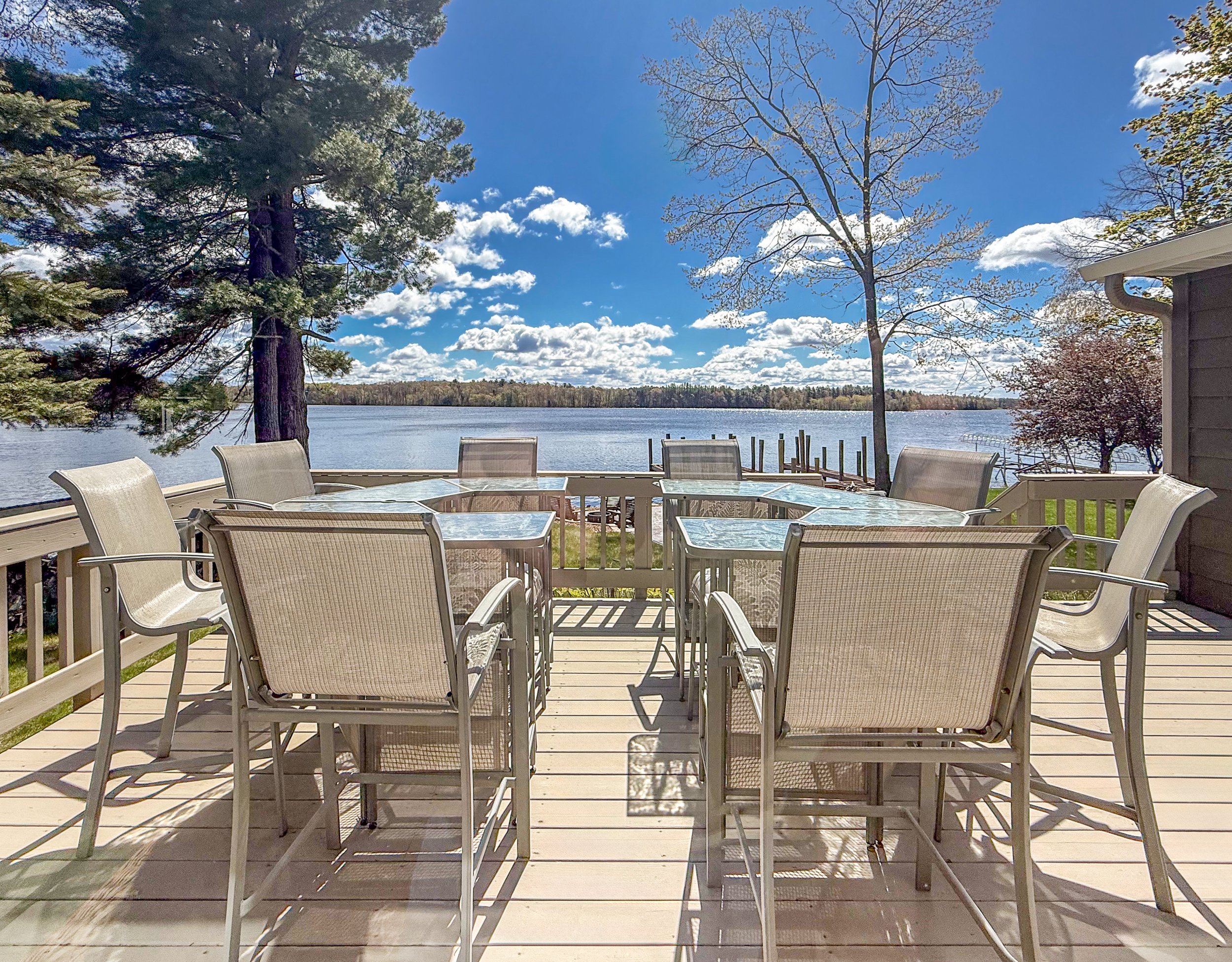 Outdoor deck with dining table and chairs overlooking a lake, with trees and a blue sky with clouds.