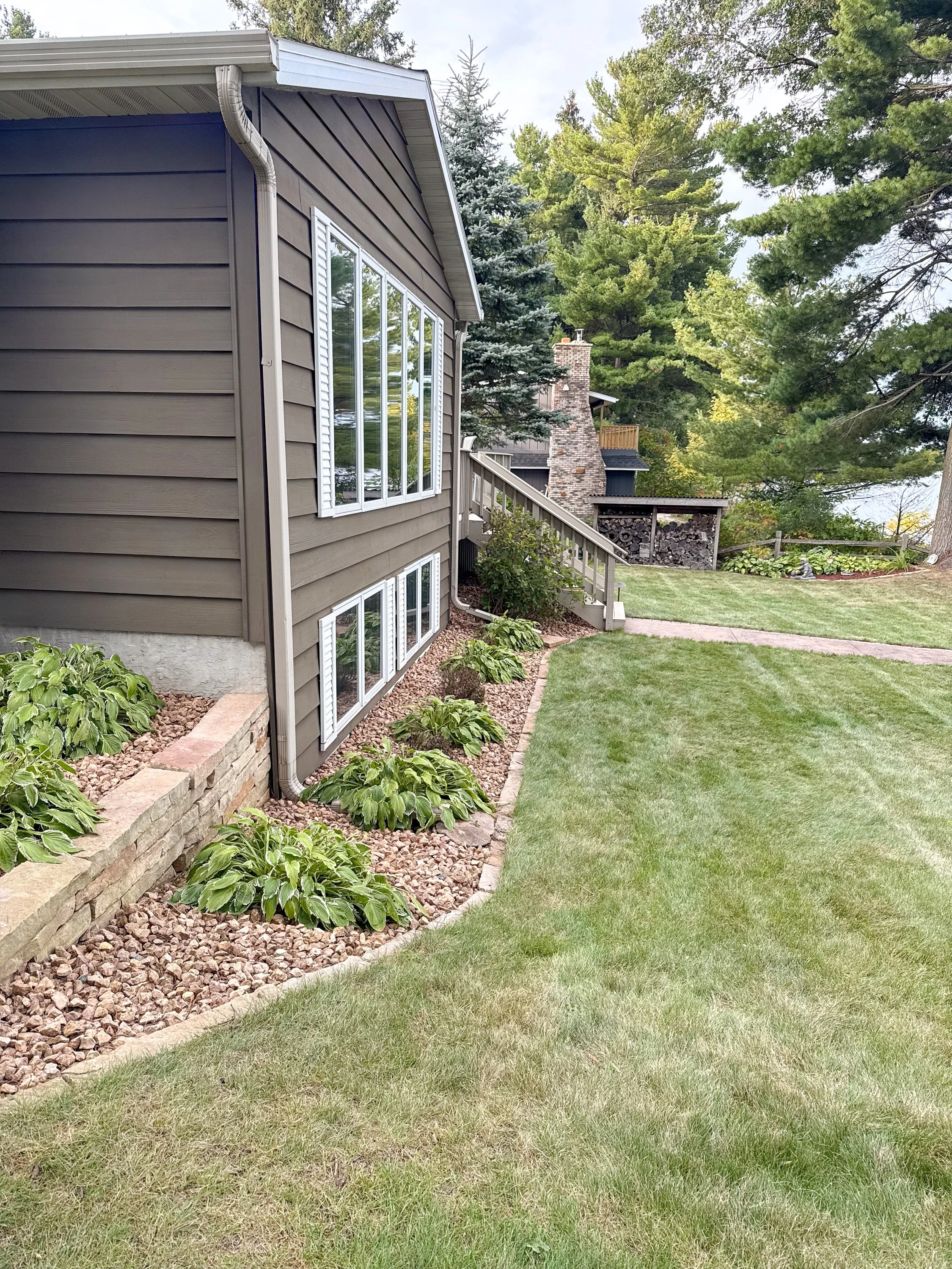 A backyard with a green lawn, landscaped garden bed with plants, a stone border, and a house with siding and large windows, surrounded by tall trees.